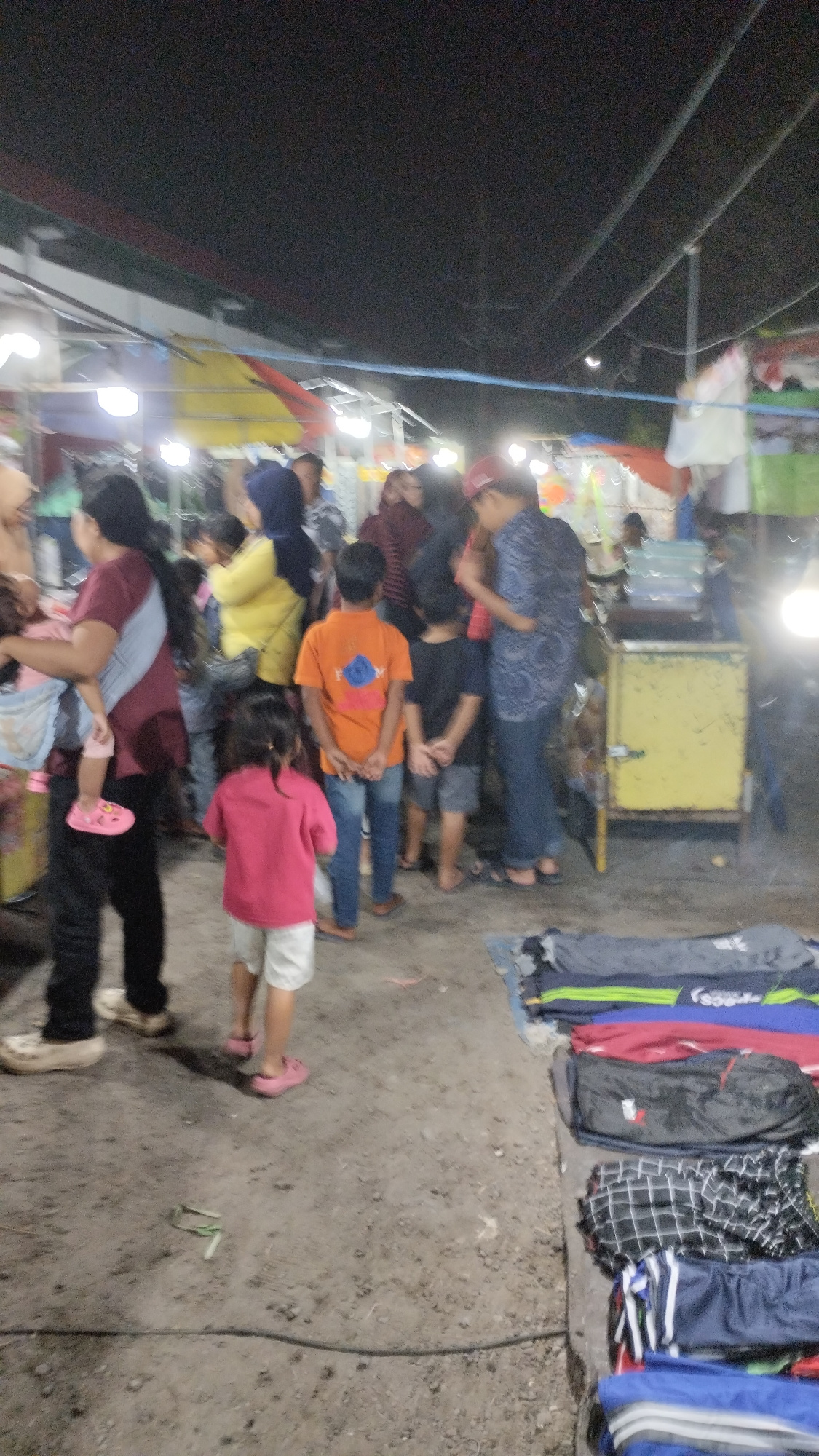 This media file depicts a nighttime scene at what appears to be a market or street fair in Sambungrejo, Indonesia. The setting is outdoors, with artificial lighting illuminating the area. Various individuals, including adults and children, are present, suggesting a public gathering.
The subjects include a woman carrying a child, a child in a pink shirt and white shorts, a boy in an orange shirt and blue jeans, and several other people of varying ages. Objects visible include market stalls with canopies, a yellow cart or kiosk, and folded garments laid out on a tarp or mat, possibly for sale. The ground appears to be dirt or gravel.
The activity taking place seems to be shopping or socializing at the market. The time of day is clearly night, indicated by the dark sky and artificial lighting. The weather is not discernible from the image. There is no visible text in the image. The general mood appears casual and bustling, typical of a night market environment.
