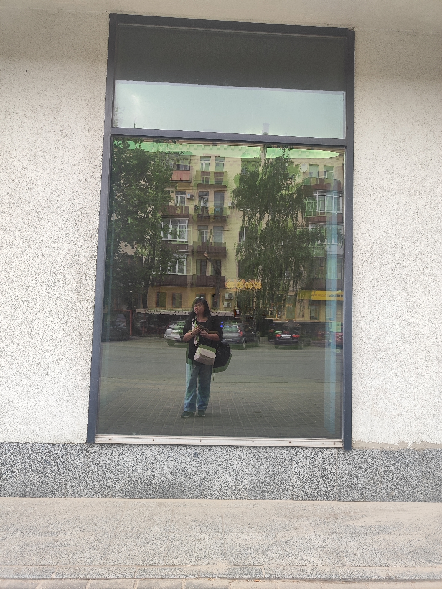 This media file shows a reflection in a large window of a building. The reflection shows a person standing in front of the window, holding a bag and a phone. The person appears to be female, with dark hair and glasses, wearing a dark top and blue jeans. She is looking at the phone. Behind her, the reflection shows a street scene with trees, buildings, and cars. The buildings appear to be apartment buildings with multiple stories and balconies. The sky in the reflection is a pale gray, suggesting an overcast day. The person's emotion is neutral, focused on her phone. The location is urban, likely a street in a city. There is some visible text on the storefronts in the reflection, but it is illegible. The overall scene is mundane and everyday.