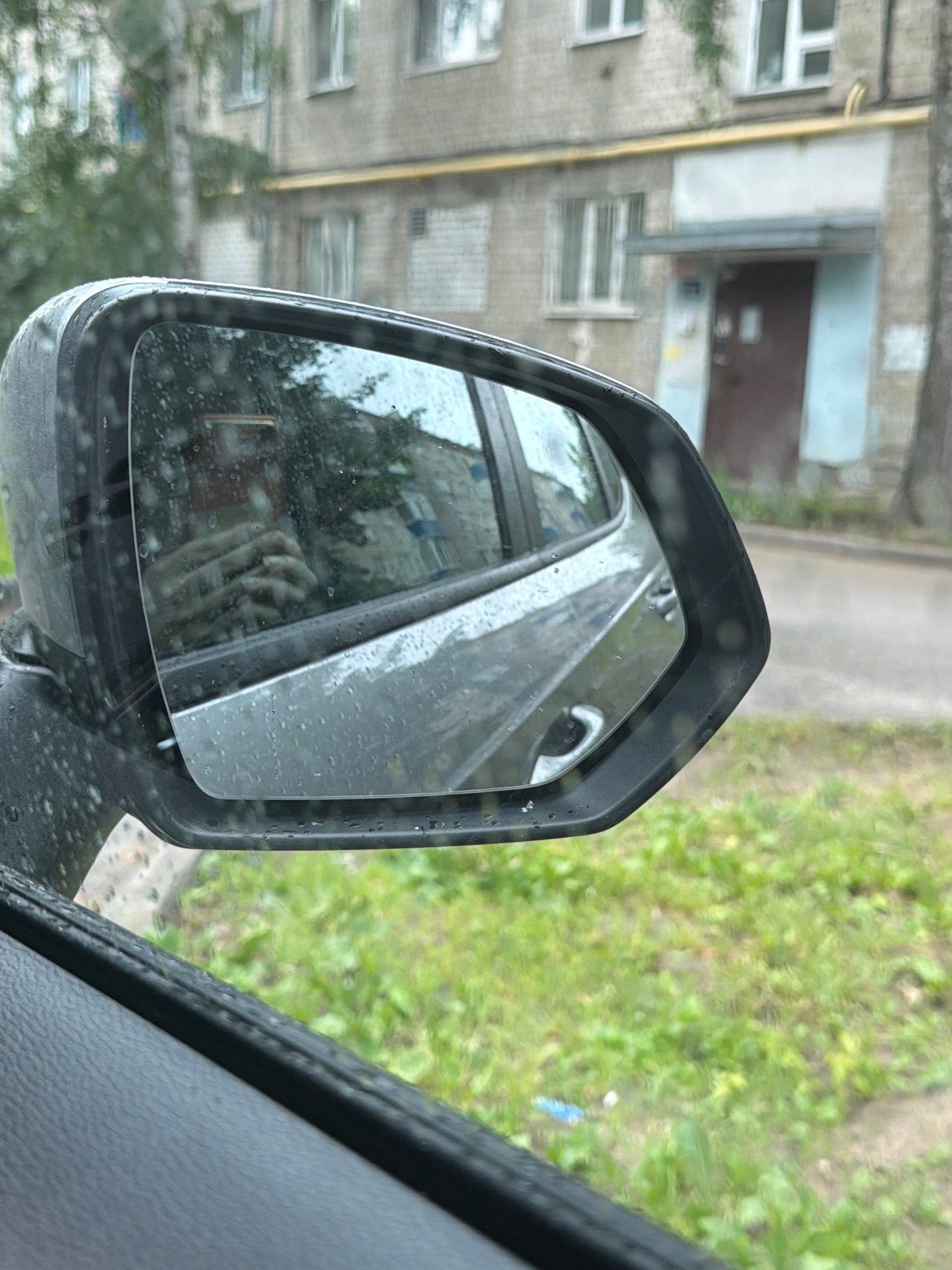 The image is a close-up shot of a car's side-view mirror, showing a reflection of the car's interior and the exterior environment. The mirror is wet with raindrops, creating a slightly distorted view. In the reflection, a person's hand holding a smartphone is visible, suggesting the photo was taken by the driver. The exterior reflection shows a grey building with multiple windows and a brown door, likely an apartment building. The environment appears to be overcast or rainy, as indicated by the raindrops on the mirror and the wet appearance of the building and the green grass in the foreground. The scene is set in an urban environment, possibly a residential area. The specific location in Kazan, Russia cannot be determined from the image alone.