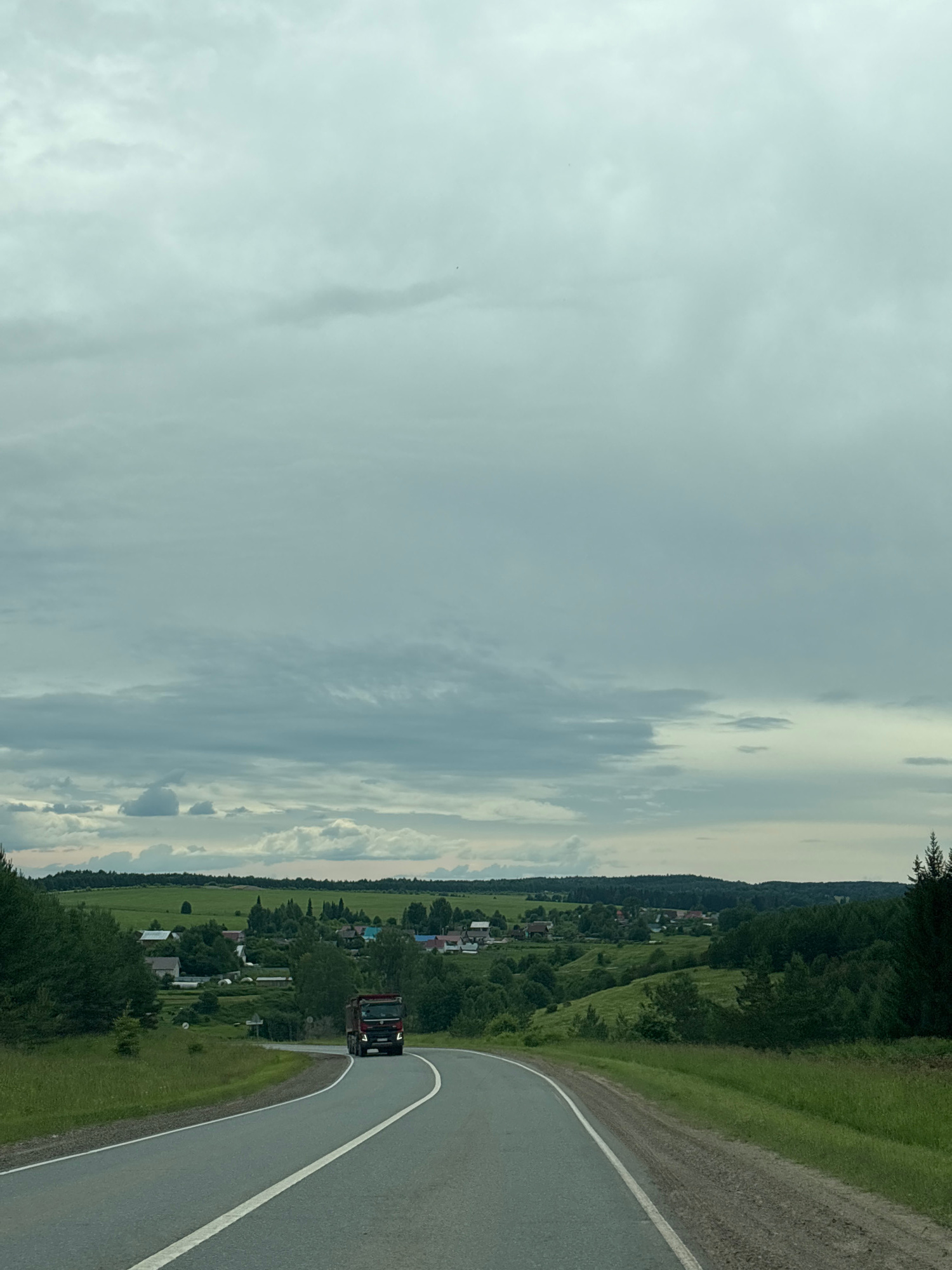 The image depicts a rural scene in Забегалово, Russia. A red truck is driving on a paved road that curves to the right. The road is bordered by a grassy embankment on the left and a dense green forest on the right. Beyond the embankment, a village is nestled in a valley. Houses with red and blue roofs are visible amidst lush green fields and trees. The sky is overcast with scattered clouds, suggesting a partly cloudy or grey day. The time of day appears to be daylight, possibly late afternoon or early evening given the lighting. The overall mood is serene and peaceful, capturing a typical rural landscape. There is no visible text in the image.