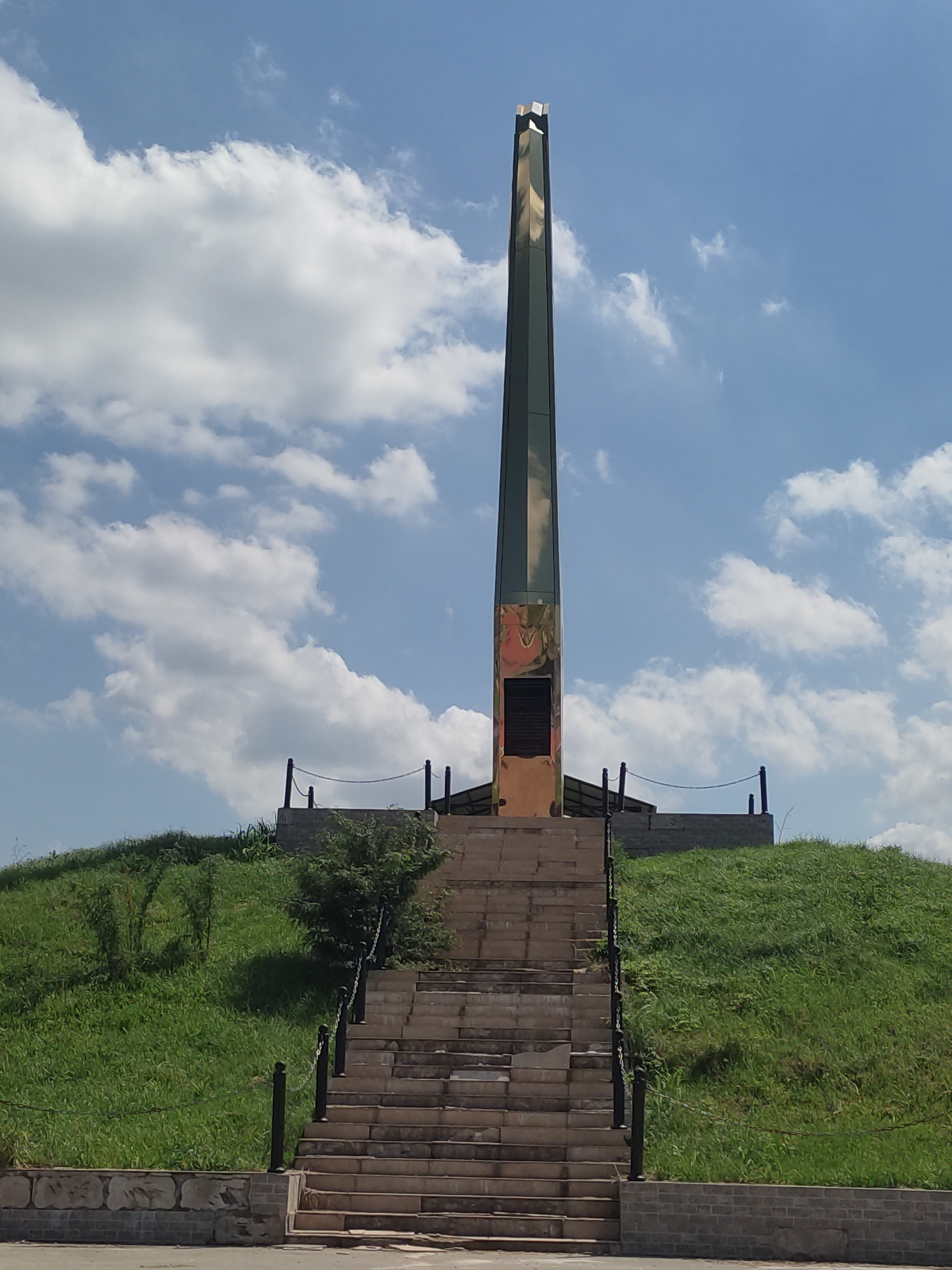 The image shows a tall, slender monument with a pointed top, positioned on a grassy hill. The monument appears to be made of metal and has some colorful imagery or markings on its lower section. A wide stone staircase leads up the hill towards the monument, with metal posts and chains along the sides. The hill is covered in lush green grass and some scattered bushes. The sky above is blue with scattered white clouds. The overall scene suggests a memorial or monument site in an outdoor location. The monument is the Memorial to the Soldiers of the Chechen War in Starye Atagi, Russia.
