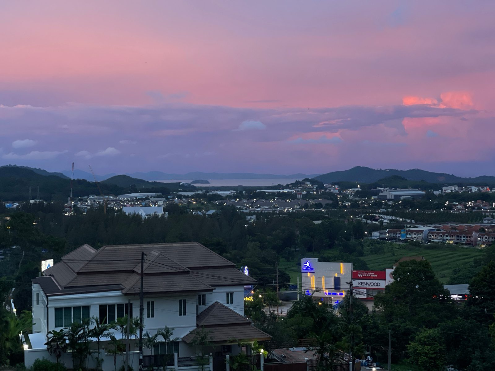 This is a wide-angle, eye-level photograph capturing a twilight scene overlooking a coastal town in Ratsada, Thailand. The sky is a gradient of soft pinks and purples, transitioning into a muted blue, with wispy clouds suggesting the last moments of sunset. In the foreground, a large, modern white house with a brown tiled roof is partially visible, with lush green foliage and palm trees surrounding it.
Beyond the house, the landscape unfolds into a sprawling town dotted with buildings, interspersed with dark green trees and patches of cultivated land, possibly rice paddies, that appear as textured green stripes. In the distance, a body of water, likely the Andaman Sea, is visible, bordered by hills and mountains that fade into the dusky horizon. The lights of the town are beginning to twinkle, indicating the transition from day to night.
Notable details include two prominent commercial buildings with illuminated signage. One building displays a blue logo with an athletic figure and the text "AIRPORT" above it. Adjacent to this is a red and white sign for "CARRYBOY" and "KENWOOD". The presence of a distant construction crane on the left side of the image suggests ongoing development in the area. The overall atmosphere is serene and tranquil, evoking a sense of calm as the day draws to a close. The time of day is clearly twilight, with the soft, diffused light characteristic of this period.