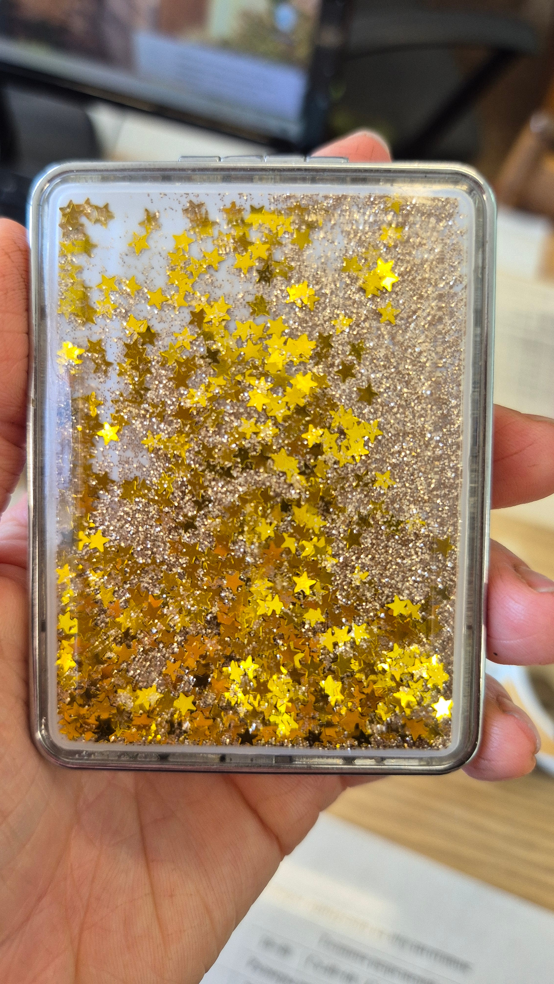 A clear, rectangular case filled with liquid and glitter is being held in a person's hand. The liquid contains numerous small, gold, star-shaped glitter pieces and finer gold glitter. The case has a metallic silver trim. The background is blurred but appears to be an indoor setting with a surface that looks like a desk or table. There is no visible indication of the city of Sterlitamak, Russia in this image.