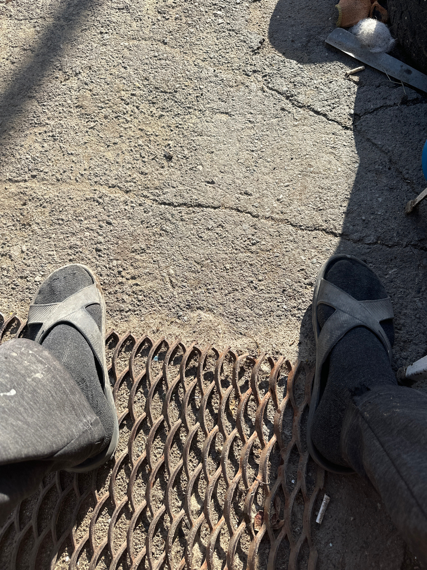 The image shows a ground-level view of two feet wearing dark gray socks and black flip-flops. One foot is positioned on a cracked concrete surface, partially covered by a shadow. The other foot is stepping onto a rusted metal grate with a diamond-shaped pattern, which rests on the concrete. The grate casts shadows on the ground. To the left of the concrete surface, there are some discarded items, including a piece of metal, a white fluffy material, and a brown fabric. A cigarette butt is visible on the concrete next to the grate and the upper foot. The lighting suggests it is daytime with the sun casting strong shadows. The scene appears to be outdoors in a somewhat neglected or industrial area, possibly a sidewalk or an alley. There is no visible text in the image.