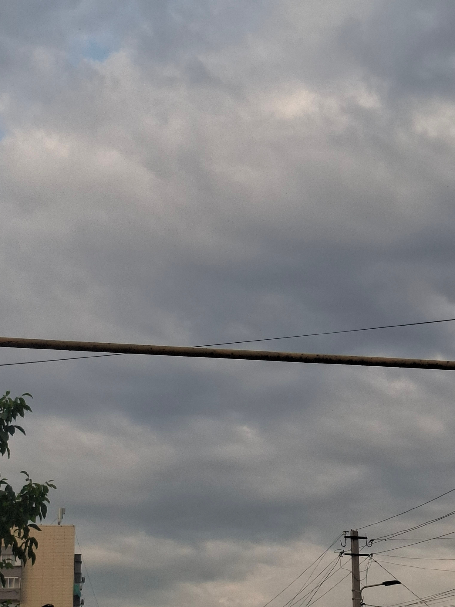 The image depicts an overcast sky with dramatic cloud formations. A prominent vertical utility pole stands in the foreground, with a thin wire extending diagonally across the frame. To the right, another utility pole is visible, adorned with numerous wires and a black fixture that appears to be a light. In the lower right corner, a portion of a building is visible, suggesting an urban or suburban setting. The building has a light-colored facade and dark elements on its roof, possibly water tanks or air conditioning units. Dark green foliage from trees partially obscures the lower part of the building, adding a touch of nature to the scene. The overall lighting and cloud cover suggest it is either early morning or late afternoon, under a cloudy or potentially stormy sky. The image appears to be taken outdoors in Almaty, Kazakhstan. No visible text is present in the image.