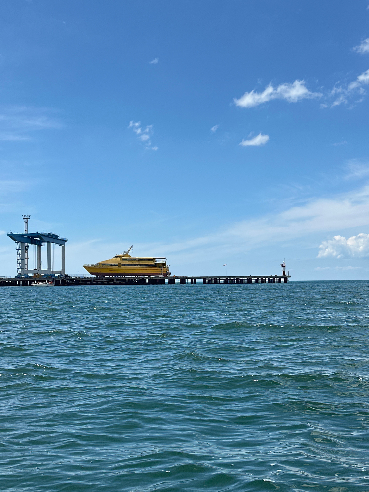 The image depicts a bright, sunny day with a clear blue sky and scattered white clouds over a body of water. In the foreground, the water is choppy with visible ripples and some small whitecaps, suggesting a moderate breeze. A long, dark pier extends from the left side of the frame towards the right, supported by numerous pilings.
On the pier, a striking bright yellow vessel, possibly a ferry or a specialized ship, is positioned. To the left of the yellow vessel, there is a large, industrial-looking structure, likely a gantry crane, with blue and grey components. Further along the pier to the right, a small red and white lighthouse or navigational marker is visible. The pier itself appears to be constructed of concrete and metal.
The scene is likely located at a port or shipyard in Anapa, Russia, given the presence of maritime infrastructure and vessels. There are no people clearly visible in the image. The overall impression is one of maritime activity or construction in a coastal setting.