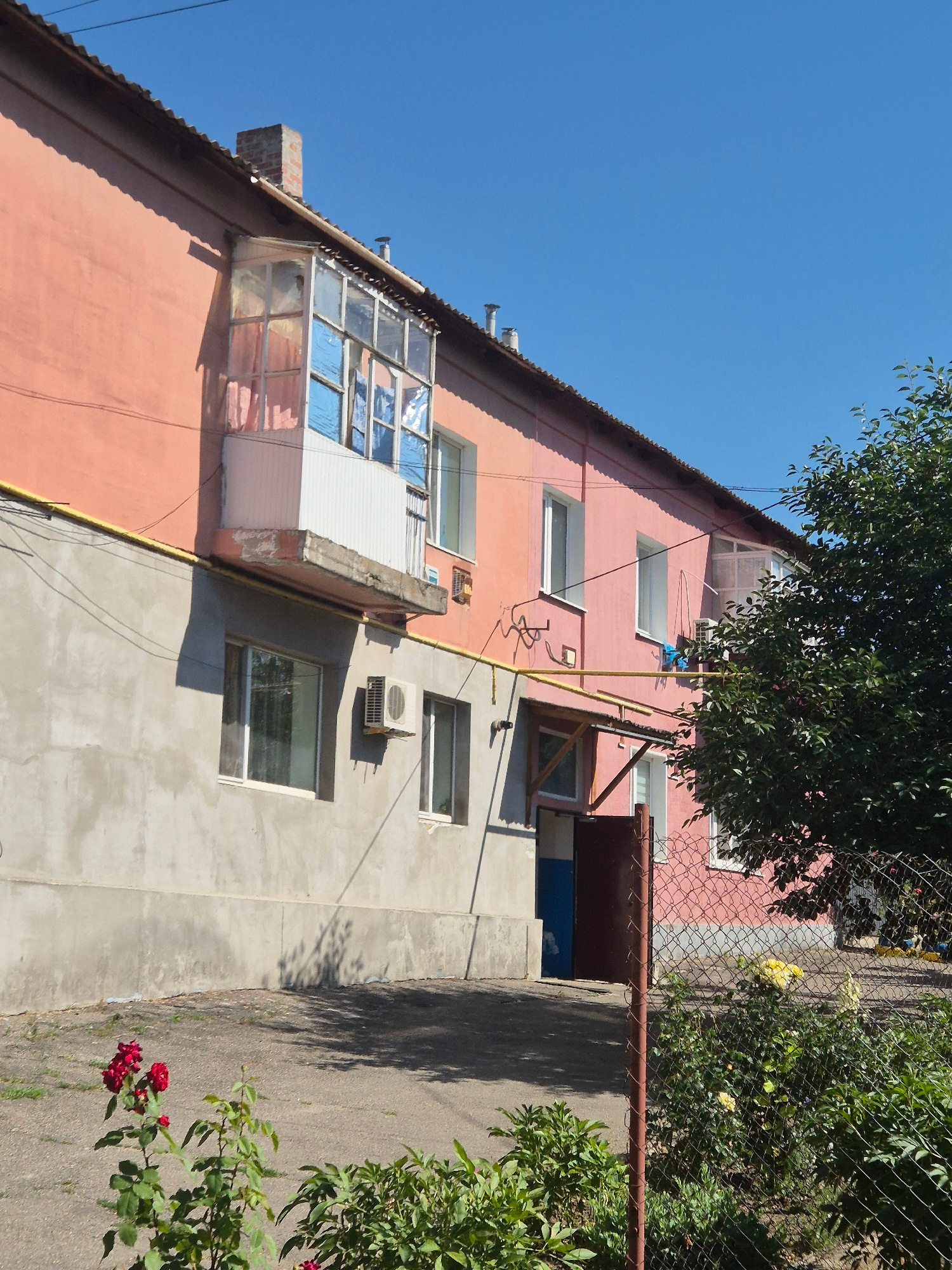 This media file is a photograph of a pink apartment building on a sunny day. The building has several windows, a balcony, and an air conditioning unit. There are trees and a fence visible in the background. The scene appears to be in a residential area, likely in Sozonivka, Ukraine, given the location context. The time of day is daytime, indicated by the bright sunlight and shadows. The weather appears to be clear and warm. There are no people or specific activities depicted in the image. The focus is on the exterior of the building and its surroundings.