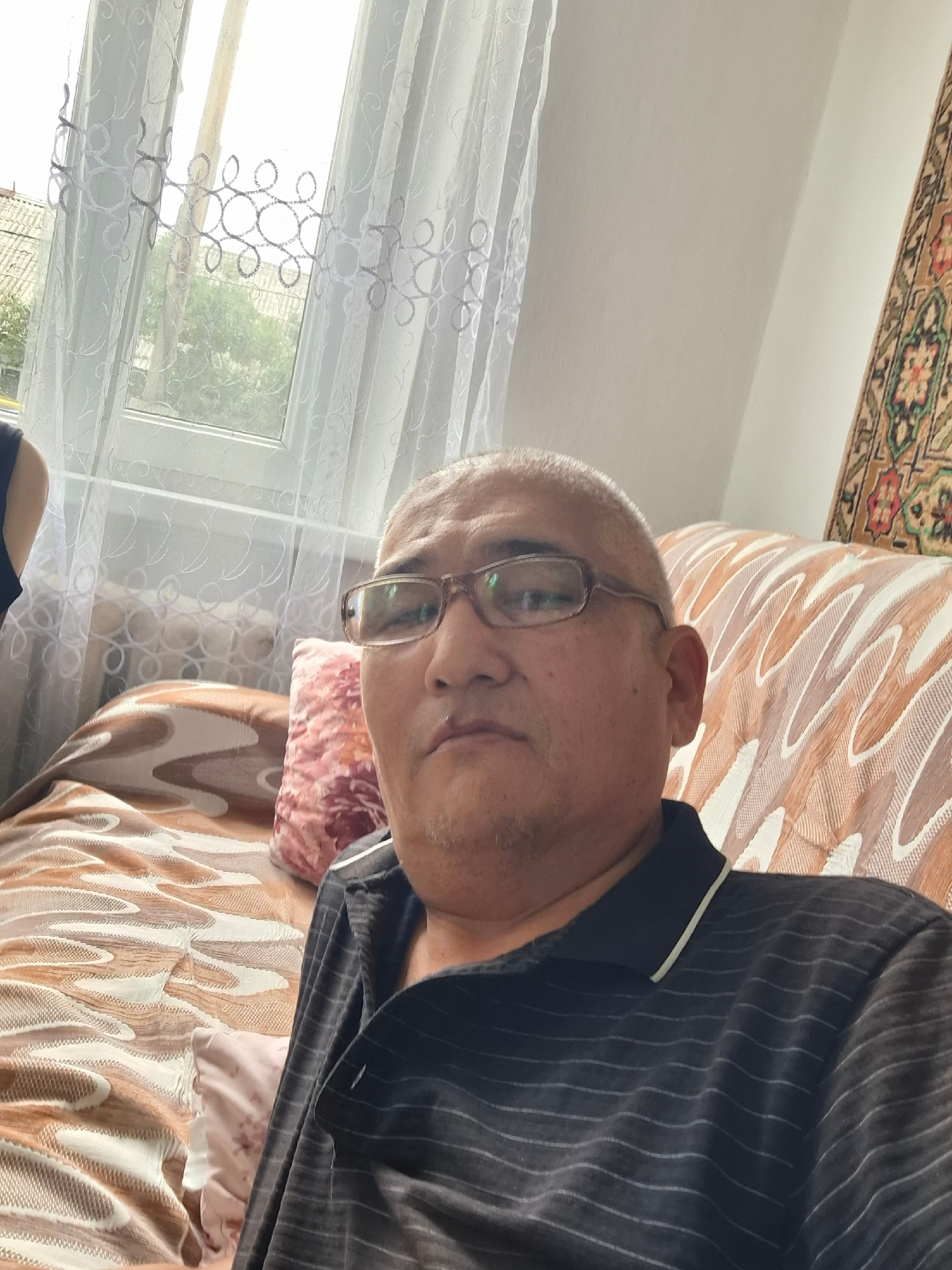 The image shows a man lying on a patterned couch. He is wearing a dark pinstripe shirt and glasses. He appears to be looking upwards, with his head resting on the couch. A window with sheer curtains is visible in the background, suggesting it is daytime. The couch has a repeating abstract pattern in shades of brown and cream, and a small patterned pillow is visible on it. A portion of a rug with floral patterns is visible at the bottom of the image. The overall scene suggests a relaxed indoor setting. The man's expression is neutral. There is no visible text in the image. The location is Almaty, Kazakhstan.