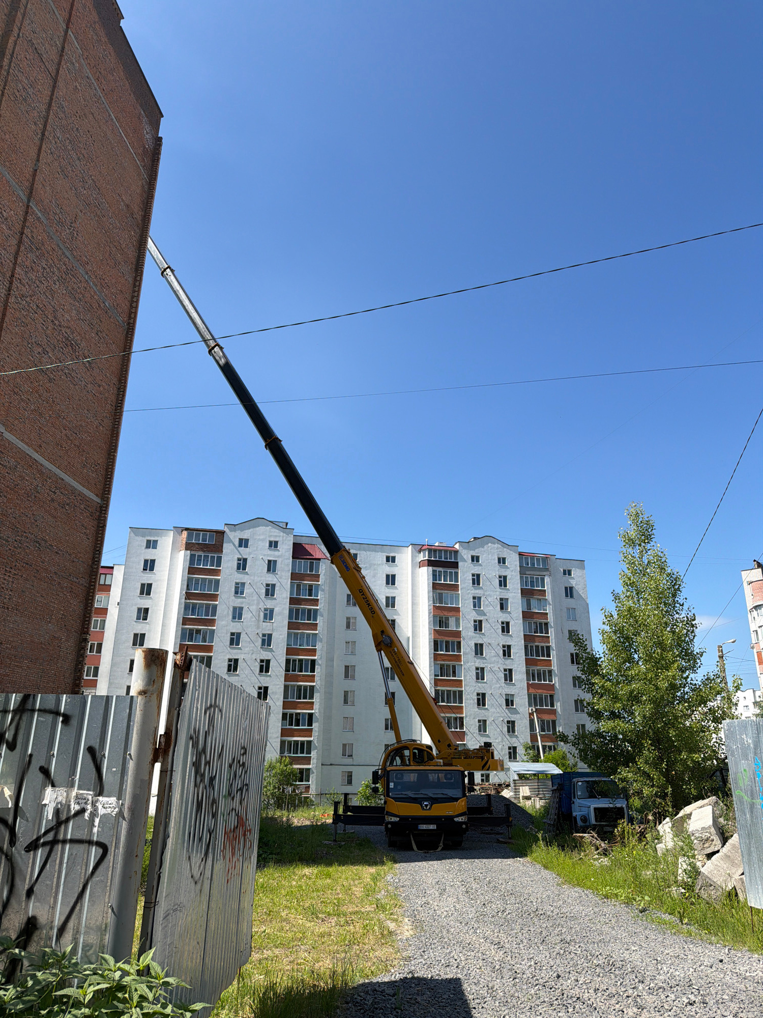 An outdoor, low-angle, full shot captures a bright, sunny day in Khmelnytskyi, Ukraine, featuring construction and urban elements. The foreground is dominated by a chain-link fence covered in graffiti. Behind it, a gravel path leads towards the center of the frame. To the left, a tall, red brick building stands, its rough texture contrasting with the smooth facade of a white apartment building in the background. A yellow crane truck is positioned on the gravel, its long boom extended diagonally across the sky, angled towards the brick building. Adjacent to the crane, a blue truck is parked, partially obscured by overgrown green grass and weeds. Several large, grey concrete blocks are piled to the right of the blue truck. A mature green tree stands between the apartment building and the concrete blocks, its leaves providing a splash of color. Overhead, a clear blue sky is crisscrossed by thin black power lines. The scene suggests ongoing construction or demolition work in a residential area of the city.