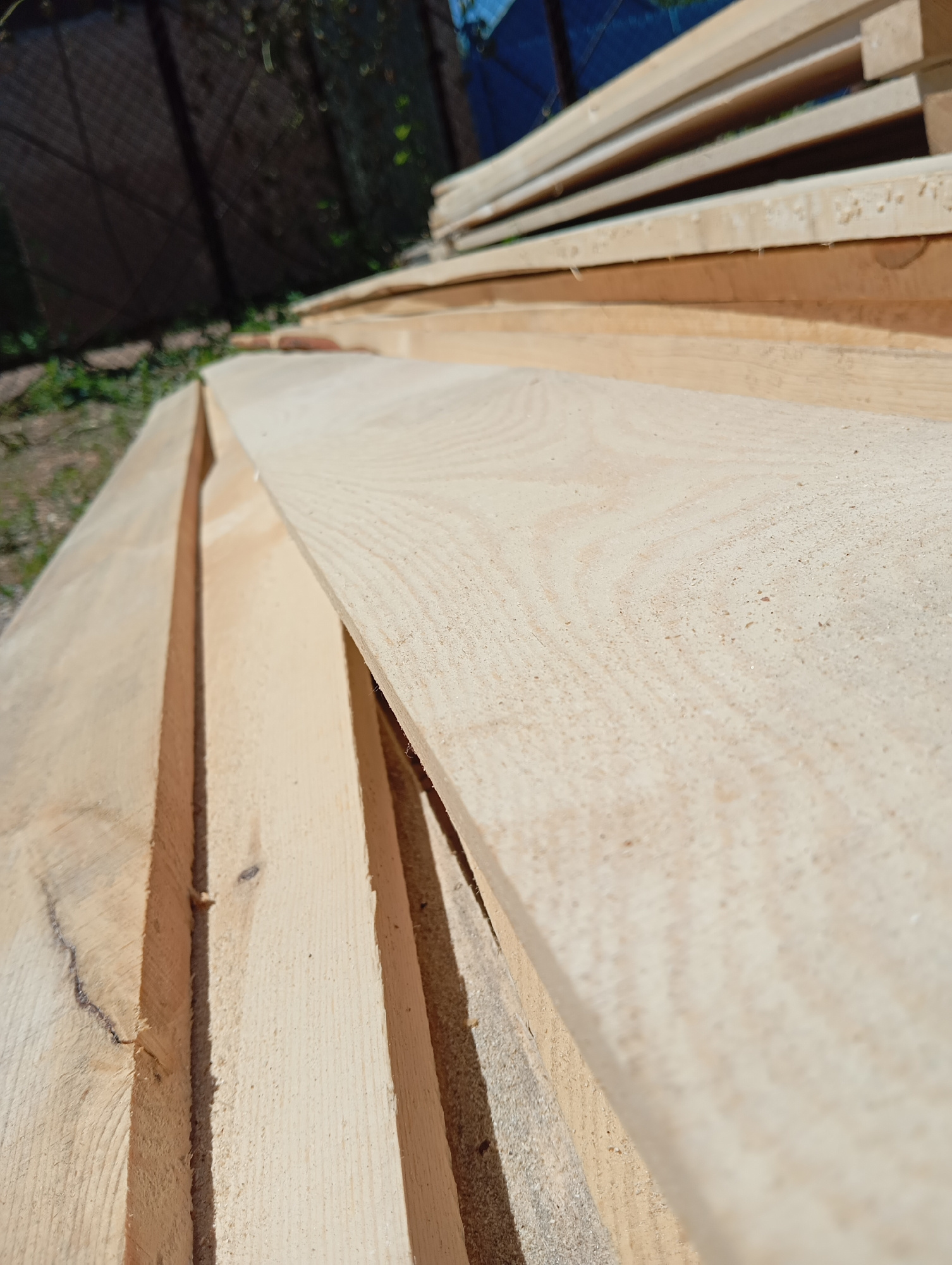 The image shows a stack of unfinished wooden planks outdoors, likely in a backyard or construction area. The planks are light-colored, with visible wood grain and some sawdust. In the background, a dark fence and some greenery are visible. The scene appears to be in a residential setting. No people or specific actions are depicted. The lighting suggests it is daytime, possibly sunny.