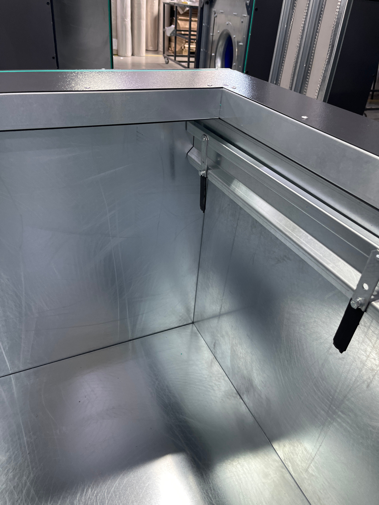 The image shows the interior of a large, metal rectangular structure, possibly a ventilation duct or container. The interior surfaces are brushed metal, showing some scuff marks and reflections. Along the top edge, a silver metal track with black rubber bumpers is visible, attached to brackets. The background is blurred but suggests an industrial or workshop environment with rolls of material and some equipment. There are no people or animals in the image. The location is not specified in the image or the prompt.