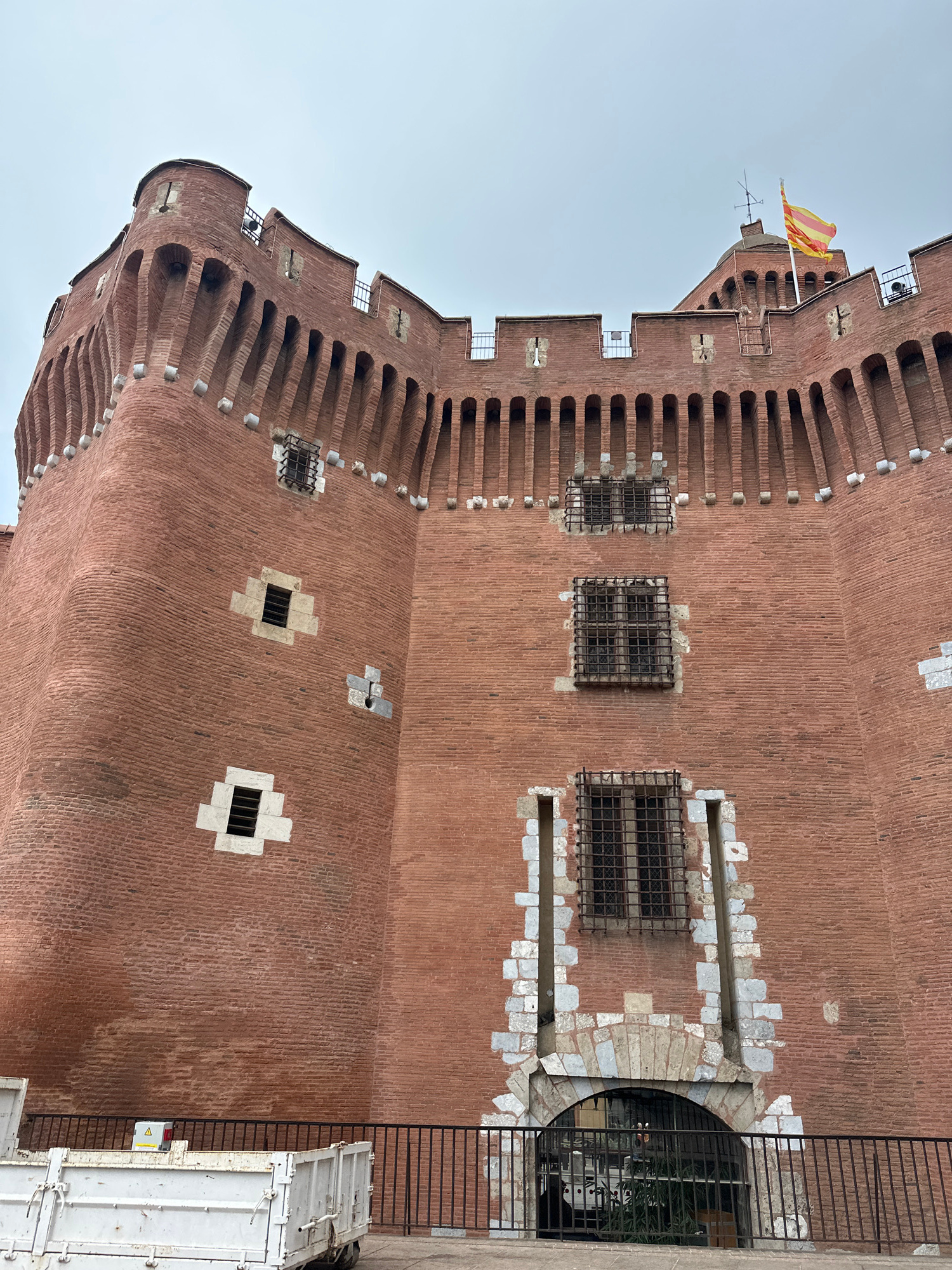 This is a vertically oriented, close-up, eye-level shot of a large, brick tower. The tower is constructed from reddish-brown bricks, with lighter-colored stone accents around the windows and along the crenellated top edge. The windows are small and some have black metal bars. A flag, which appears to be yellow and red striped, flies from a pole near the top of the tower. The sky is a pale, overcast blue, suggesting it is daytime. The scene appears to be outdoors, with a black metal fence and a white truck visible on the right side of the image. The building's architecture suggests it might be a historical structure, possibly a castle or fortified tower. The overall impression is one of solid construction and historical significance. There is a yellow triangle caution sign visible on the white truck.