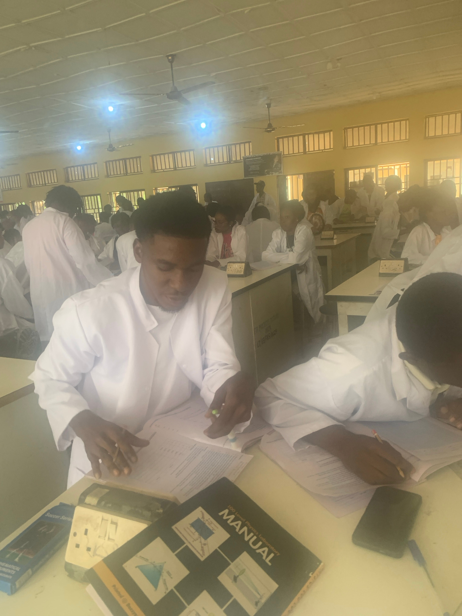 The image depicts a classroom setting where numerous individuals, primarily young adults, are seated at desks. They are all wearing white lab coats, suggesting a science or medical-related educational environment. In the foreground, a young man in a lab coat is focused on papers in front of him, holding a pen. To his right, another individual in a lab coat is also engaged with documents. Various books and papers are visible on the desks, along with a mobile phone and electrical outlets. The room has fluorescent lighting and ceiling fans, and windows with slatted blinds are present along the walls. The overall scene conveys an atmosphere of learning and academic activity.