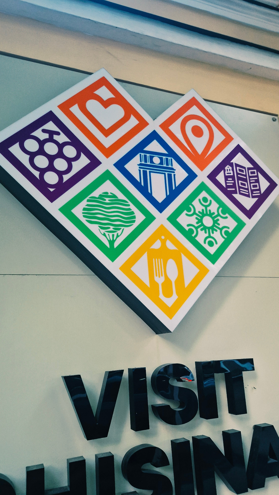 The image displays a diamond-shaped logo composed of nine smaller rhombuses, each containing a white icon on a colored background. The icons include a heart, grapes, a map marker, an archway, a tree, a snowflake-like design, and cutlery. Below the logo, large, black, three-dimensional letters spell out "VISIT" on the top line and "CHISINA" on the bottom line, suggesting the logo and signage are related to tourism for the city of Chișinău, Moldova. The signage is mounted on a light-colored wall, with a wooden ceiling element visible in the upper portion of the image.
