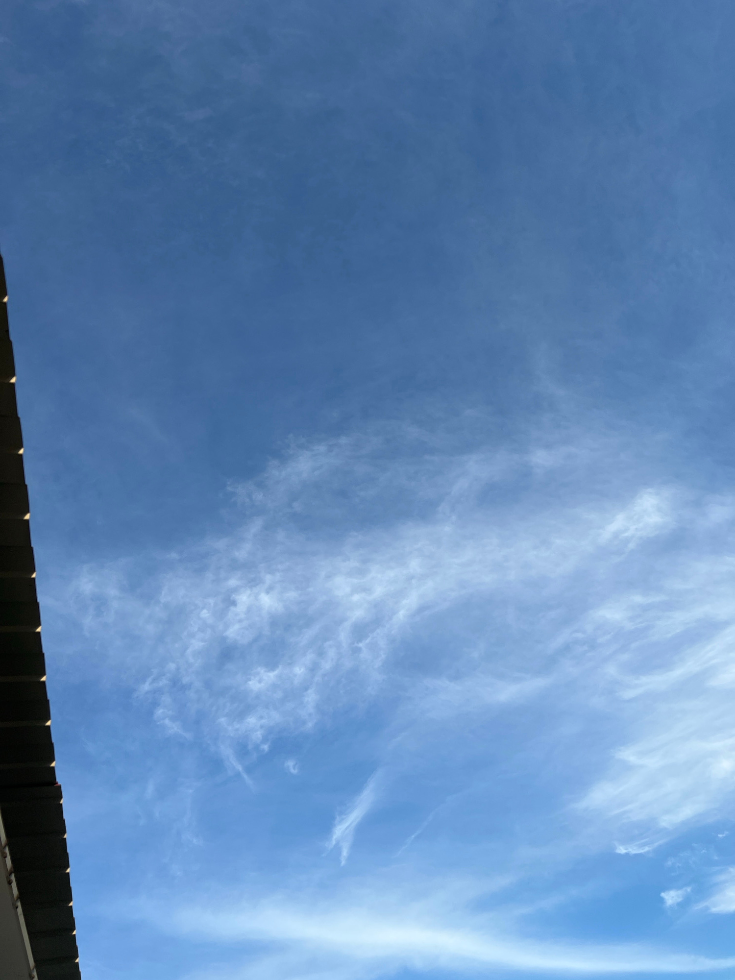 The image displays a clear blue sky with thin, wispy cirrus clouds stretching across the frame. In the lower left corner, a portion of a dark-colored roof with visible horizontal planks is visible, suggesting an outdoor scene. The image is taken from a low angle, looking upwards towards the sky. There are no people or animals present in the image. The environment appears to be a typical urban or suburban setting, possibly within the city of Verkhnesadivskyi okruh, Ukraine.