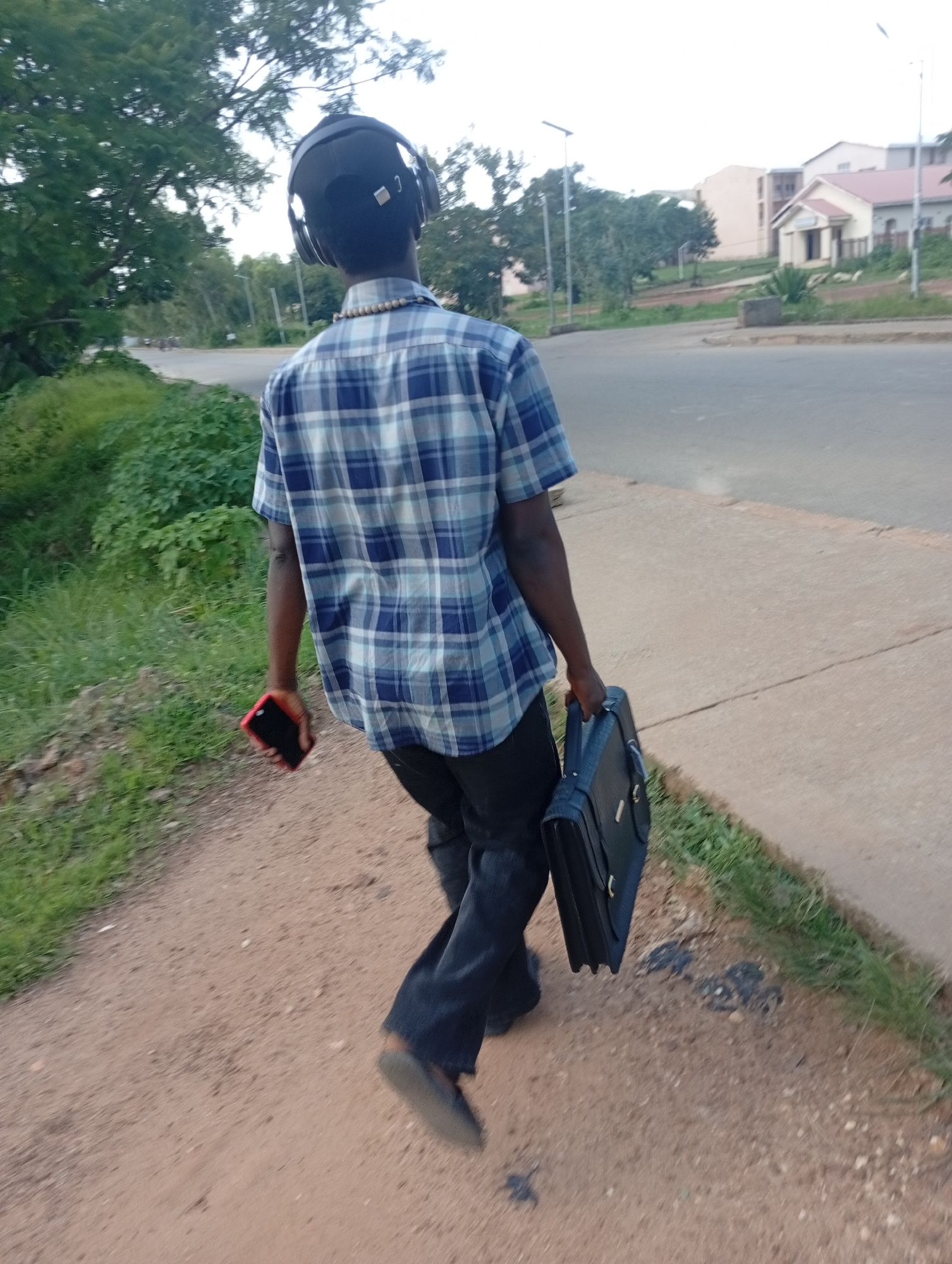 A full shot shows a Black man walking away from the camera down a dirt path next to a paved road. The man is wearing a blue and white checkered short-sleeved button-down shirt, black pants, and headphones. He is carrying a black briefcase in his right hand and a red smartphone in his left hand. In the background, there are trees, buildings, and a street. The location is Rafin Par, Nigeria.