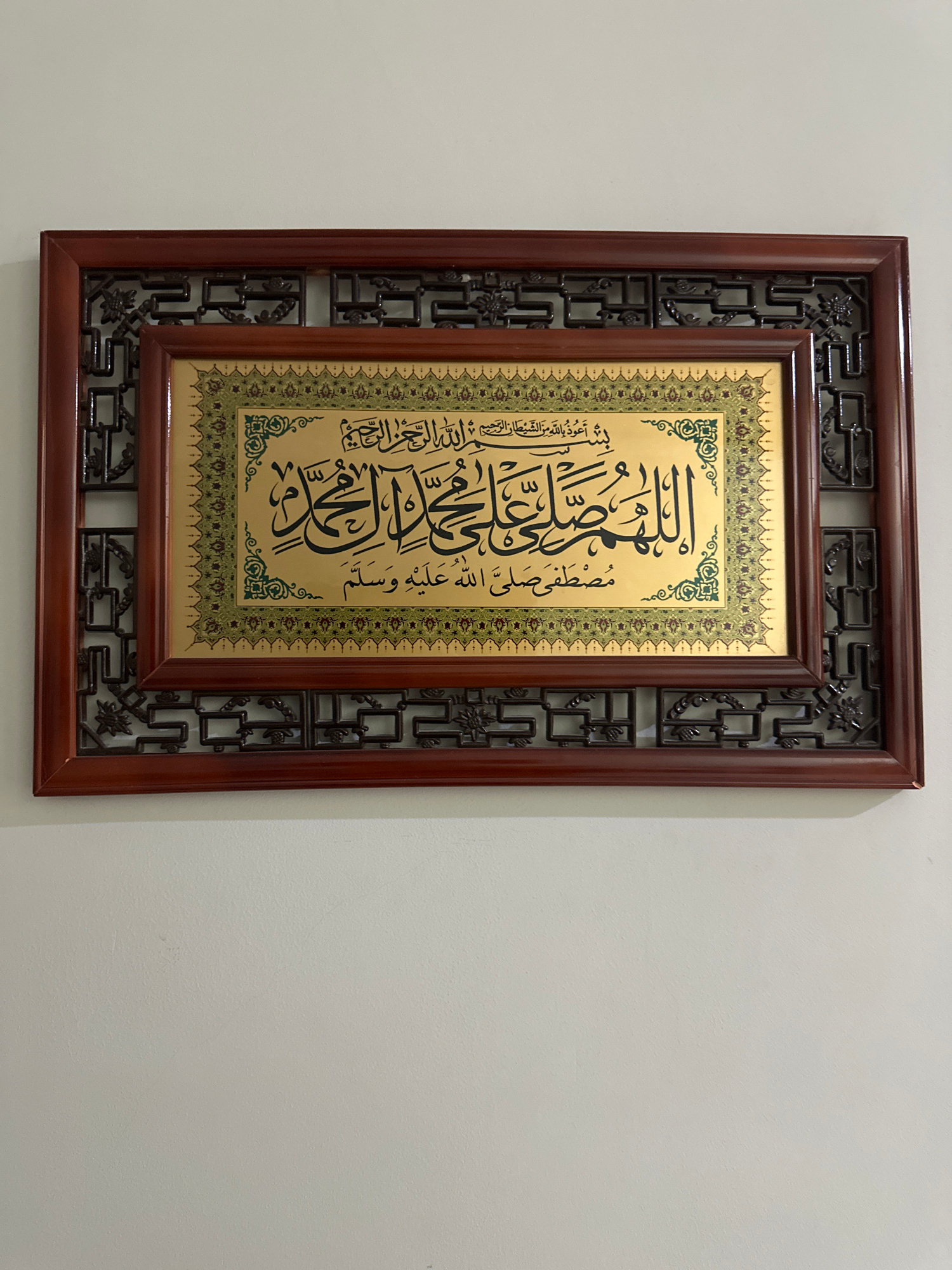 A framed artwork featuring Arabic calligraphy is mounted on a plain, off-white wall. The frame is made of dark wood with an ornate, lattice-like design along the outer edges and a smoother, inset wooden border around the central artwork. The artwork itself has a golden background with intricate green and gold patterns along the borders. The Arabic calligraphy is written in black ink, with the main text appearing large and prominent in the center. The calligraphy includes what appears to be a prayer or religious invocation, beginning with "Bismillah al-Rahman al-Rahim" at the top right. The style of the calligraphy and the decorative elements suggest it is of Islamic origin. The lighting in the scene is even, casting no harsh shadows, indicating ambient or diffused lighting. There are no people or other objects visible in the image, with the focus solely on the framed artwork and the wall it is displayed on. The environment appears to be indoors, possibly a home or a public space, with the wall providing a neutral backdrop. The image is captured from a slightly angled perspective, showing the top edge of the frame and the wall above it. There is no information in the image that would suggest a location in Tashkent, Uzbekistan.
