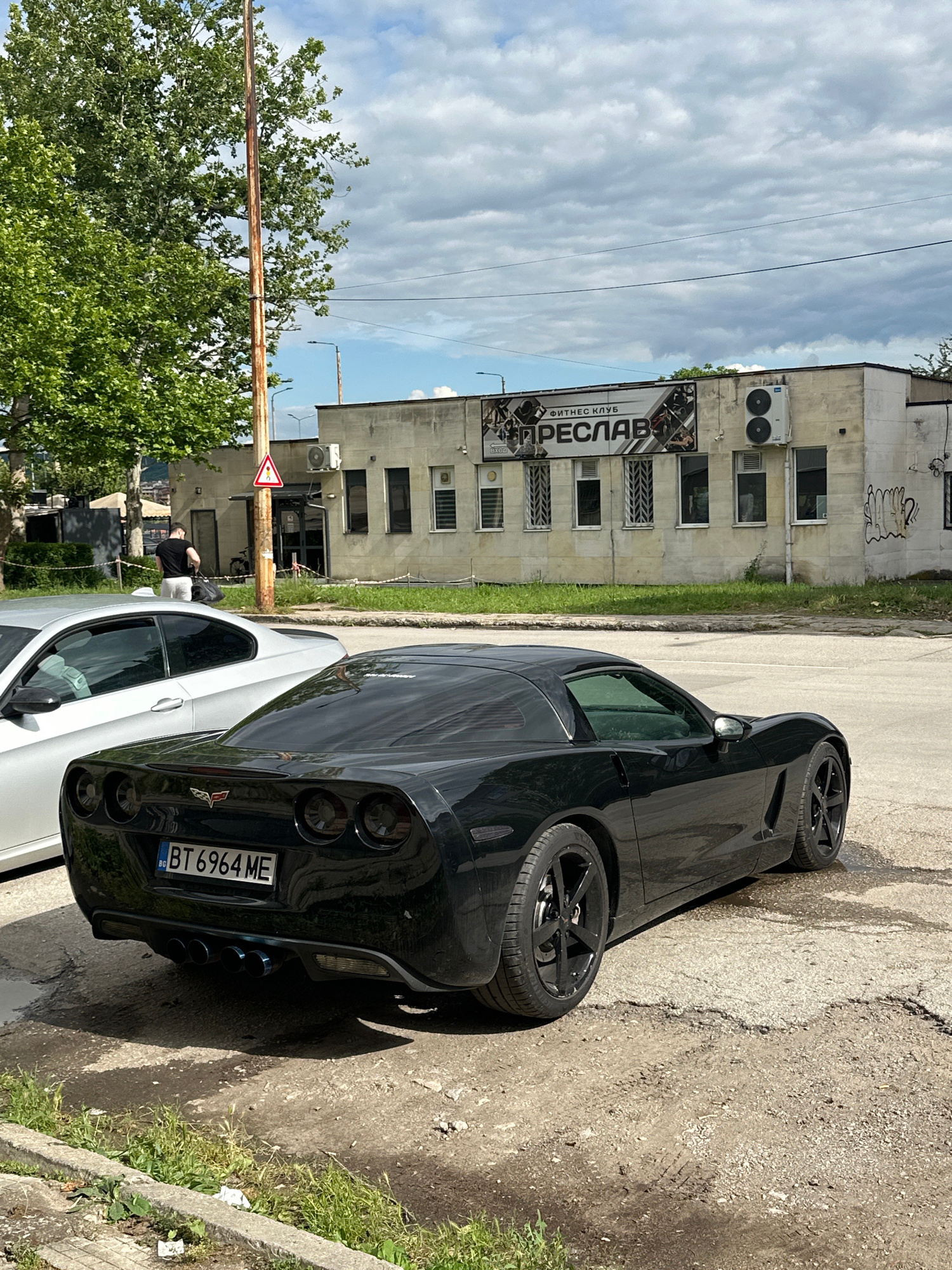 A black Chevrolet Corvette is parked on a cracked asphalt surface outside a building. The car's rear is facing the camera, and its license plate reads "BT 6964 ME". A silver car is parked to the left of the Corvette. The building behind the cars has a sign that reads "ФИТНЕС КЛУБ ПРЕСЛАВ". A man in a black t-shirt and white shorts is walking away from the building, carrying a black bag. A utility pole with a red and white triangular sign stands between the cars and the building. The scene is under a cloudy sky.