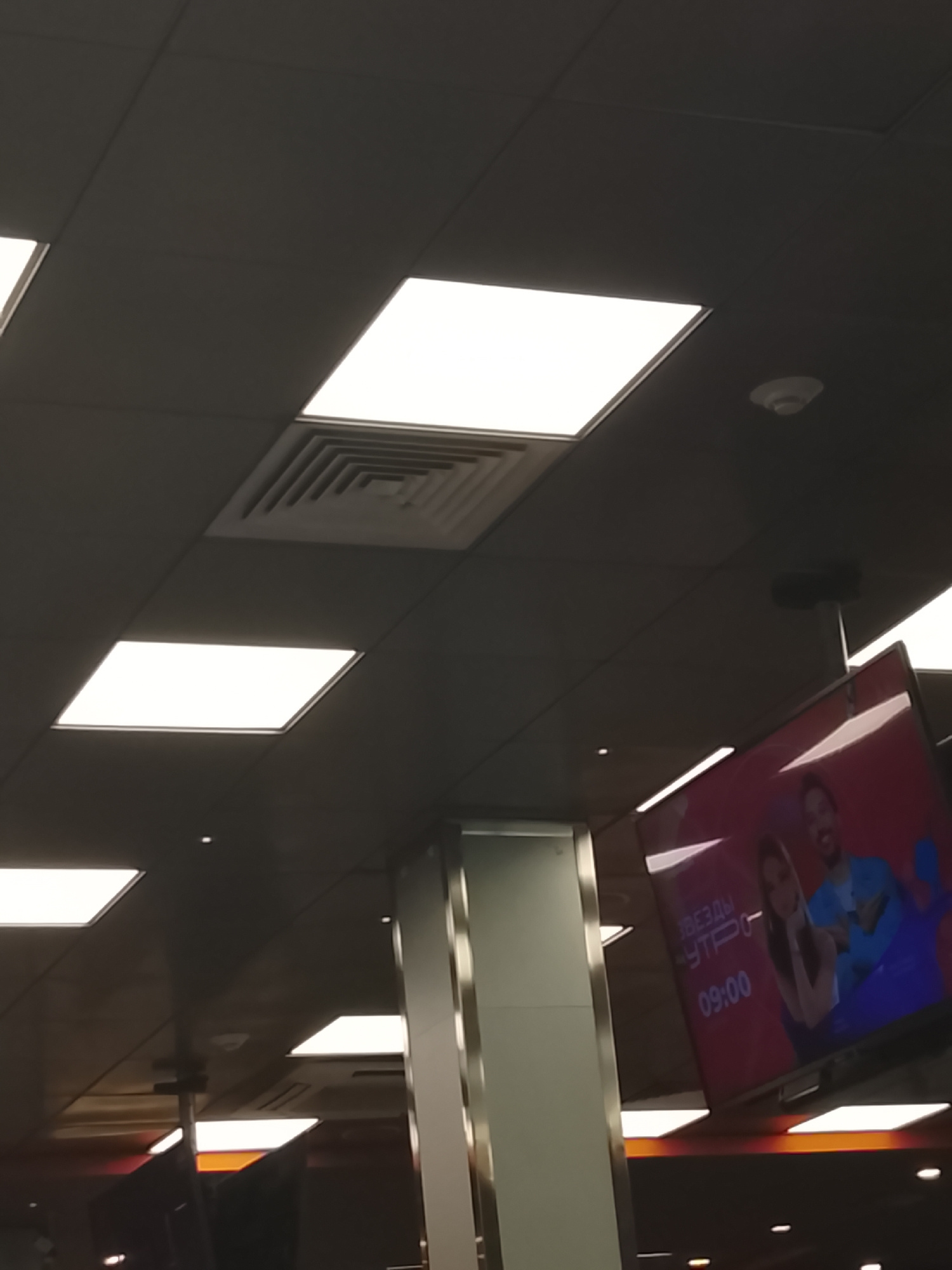The image shows a ceiling with several rectangular fluorescent light panels. In the upper right corner, a white circular smoke detector is visible. A large flat-screen television is mounted from the ceiling and displays an image of two people and text in Russian. The text on the screen reads "ЗВЕЗДЫ УТРО" and "09:00". The television appears to be showing a segment of a morning show. The overall environment suggests an indoor public space, possibly a shopping mall, airport, or other commercial establishment. The location in Almaty, Kazakhstan, is not evident from the visual information in the image.