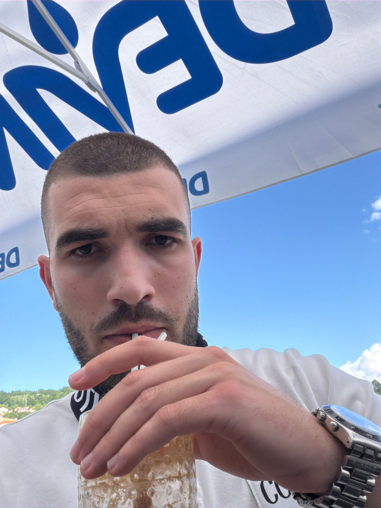 A man with a buzz cut and beard is looking at the camera while holding a drink and a straw. He is wearing a white t-shirt and a silver watch. The background is a clear blue sky with some clouds, and a white umbrella with blue lettering.
