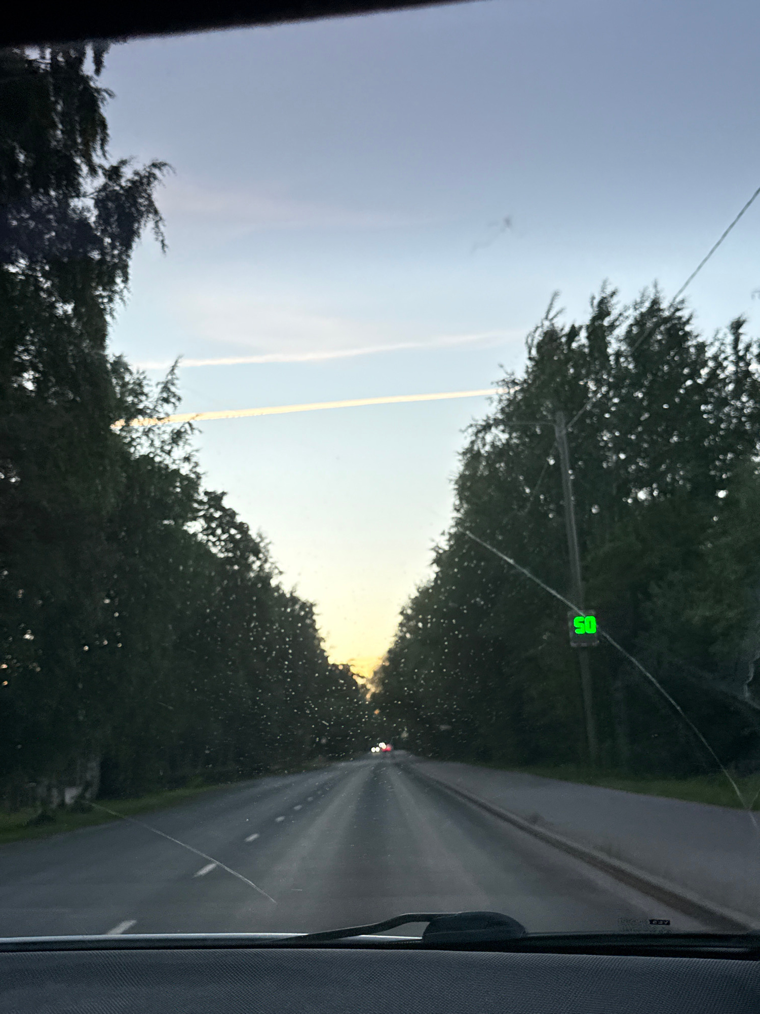The image captures a view from inside a vehicle looking forward on a road lined with trees. A speed limit sign displaying "50" in green is visible on the right side of the road. The sky is partly cloudy with a faint contrail in the distance, suggesting it is either early morning or late afternoon. The road ahead is paved and appears to be a two-lane road. There are no visible people in the image. The scene is an outdoor environment, likely a rural or suburban area given the presence of trees and the absence of significant urban infrastructure. The location is not definitively identifiable as Tallinn, Estonia, without further context or landmarks.