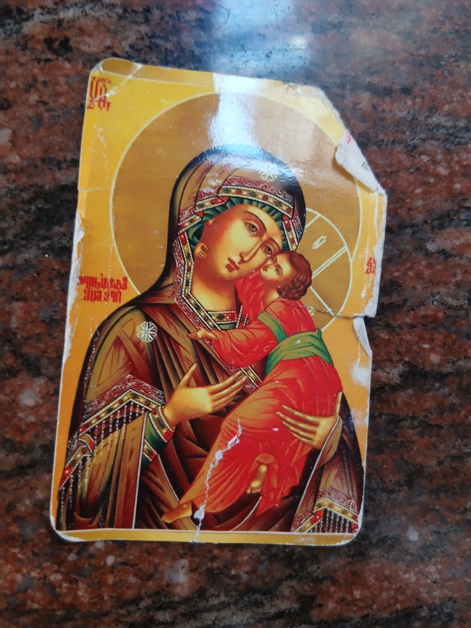 This is a close-up, slightly angled, overhead shot of a sticker depicting an icon of the Virgin Mary holding the Christ child. The sticker is yellow with gold accents and is placed on a dark, speckled surface that appears to be granite. The icon itself features rich colors, primarily reds, greens, and golds, with intricate patterns on the clothing and halos. The Virgin Mary's face is detailed, with large eyes and a serene expression. The Christ child is nestled in her arms, also depicted with a gentle expression. Both figures have halos. There is some wear and tear on the sticker, particularly along the edges and a tear across the top. Visible text in a reddish-orange color is present on the lower left and lower right portions of the sticker. The text is in a script that appears to be Armenian. The overall impression is of a religious devotional image. The setting appears to be indoors, given the lighting and the texture of the surface it's placed upon, which could be a tabletop or counter in a home or shop. The sticker is the sole focus, with the background out of focus, drawing attention to the religious imagery.