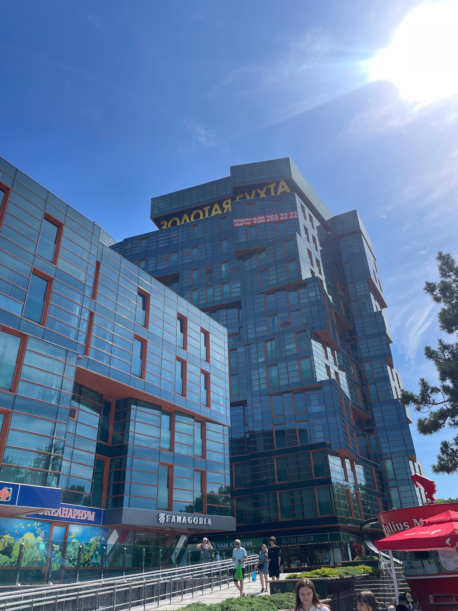 The image captures a contemporary building complex with a striking architectural design, characterized by extensive use of glass and orange-tinted window frames. The complex is illuminated by bright sunlight, which casts lens flares across the sky, indicating a clear and sunny day. The buildings appear to be multi-story, with an angular, staggered design, suggesting a modern aesthetic. One building features prominent yellow lettering that reads "ЗОЛОТАЯ ГУХТА" (Zolotaya Gukhta) and a phone number for apartment sales. The ground level of one building houses businesses, with visible signage for "АКВАНАРИУМ" (Akvanarium) and "FANAGORIA". Several people are walking along a pathway in front of the buildings, dressed casually. A red umbrella with "Julius M" visible on it suggests an outdoor seating area or kiosk. The scene is set against a bright blue sky with wispy white clouds. This building complex is located in the city of Anapa, Russia, a resort town on the Black Sea.
