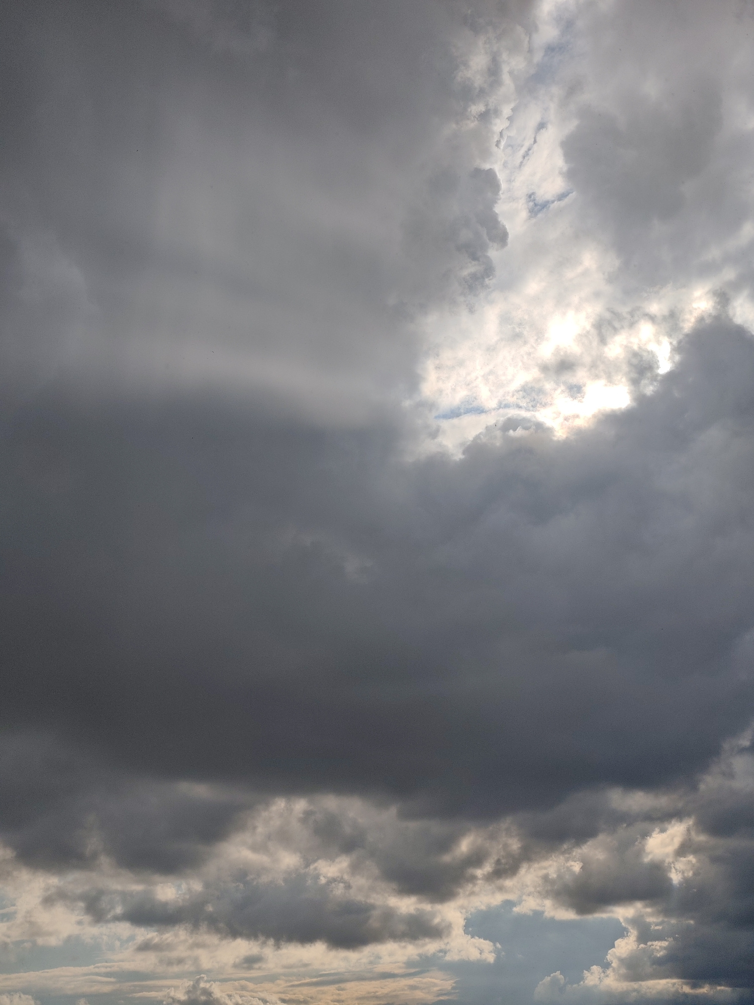 An image shows a cloudy sky. Dark gray clouds dominate the frame, with some lighter gray clouds interspersed. Sunlight breaks through the clouds in the upper right portion of the image, creating bright patches and rays of light. The sky appears to be in a state of transition, possibly before or during a storm. There are no visible people, subjects, objects, actions, or interactions. The environment is solely the sky.