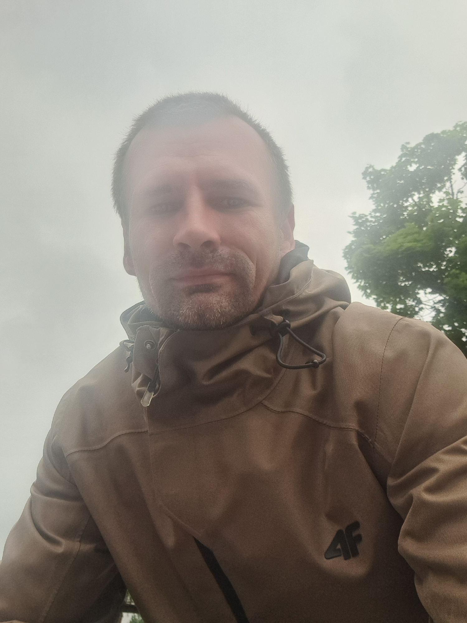 A man with short, dark hair and stubble is pictured in a close-up, low-angle selfie. He is wearing a brown hooded jacket with a black logo that reads "4F" on the chest. The background is out of focus and appears to be a foggy or overcast sky with some green foliage visible on the right side. The image quality is grainy, suggesting it was taken with a low-resolution camera or in low light conditions. The man's face is partially obscured by the angle and the focus. The image is taken outdoors, possibly in a park or natural area, given the presence of trees. No specific landmarks or identifying features of Cēsis, Latvia, are visible.