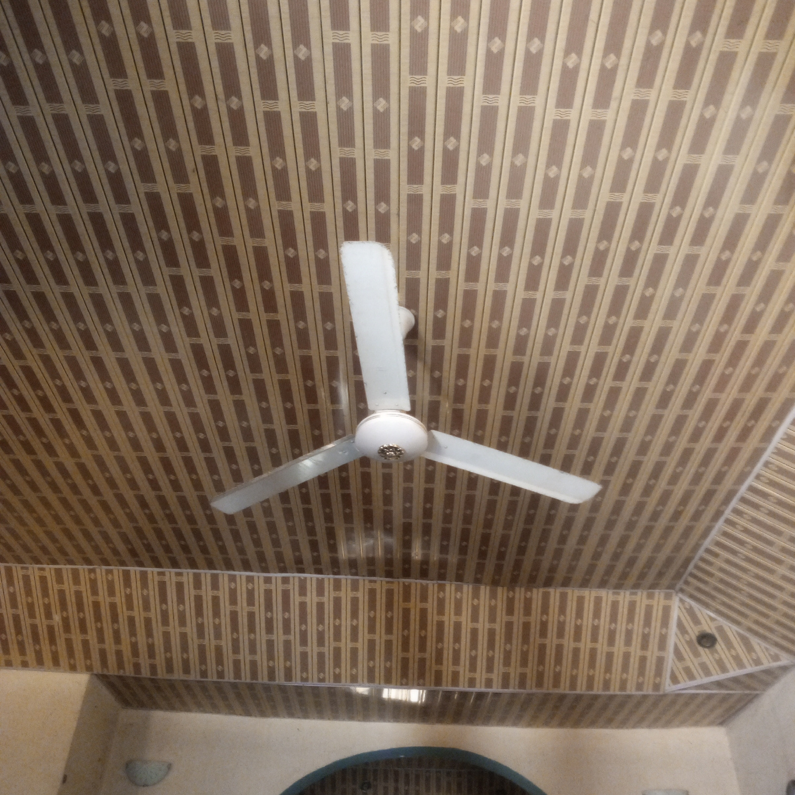 A white three-bladed ceiling fan hangs from a ceiling with a patterned design of alternating brown and beige vertical stripes, each with a small diamond shape and wavy lines within the brown sections. The fan has a white spherical central housing with a decorative gold accent in the center. One of the blades is angled upwards and to the left, another is angled downwards and to the right, and the third is angled downwards and to the left, suggesting it is either stationary or slowly rotating. The ceiling fan is located in Zinna, Nigeria.