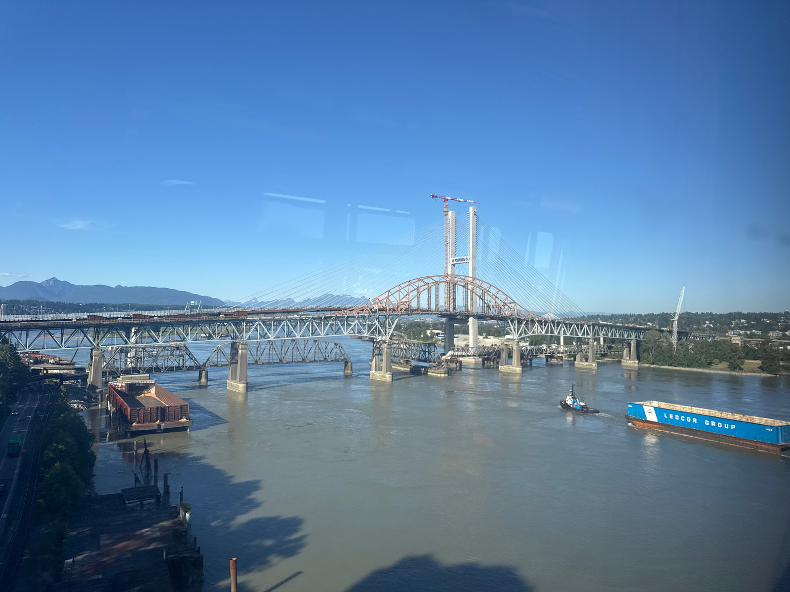 The image shows a wide, murky river flowing under a large, multi-span bridge. The bridge appears to be a combination of older truss construction and a newer cable-stayed section that is still under construction, with cranes visible at the top of its towers. On the river, a small tugboat is pushing a large, blue barge with the words "LEDCOR GROUP" printed on its side. In the distance, a range of mountains can be seen under a clear blue sky. The foreground shows a dark, partially dilapidated pier with some debris. The location is the Fraser River, adjacent to the city of New Westminster, Canada, likely in the vicinity of the Pattullo Bridge and the ongoing construction of the new Fraser River Bridge.