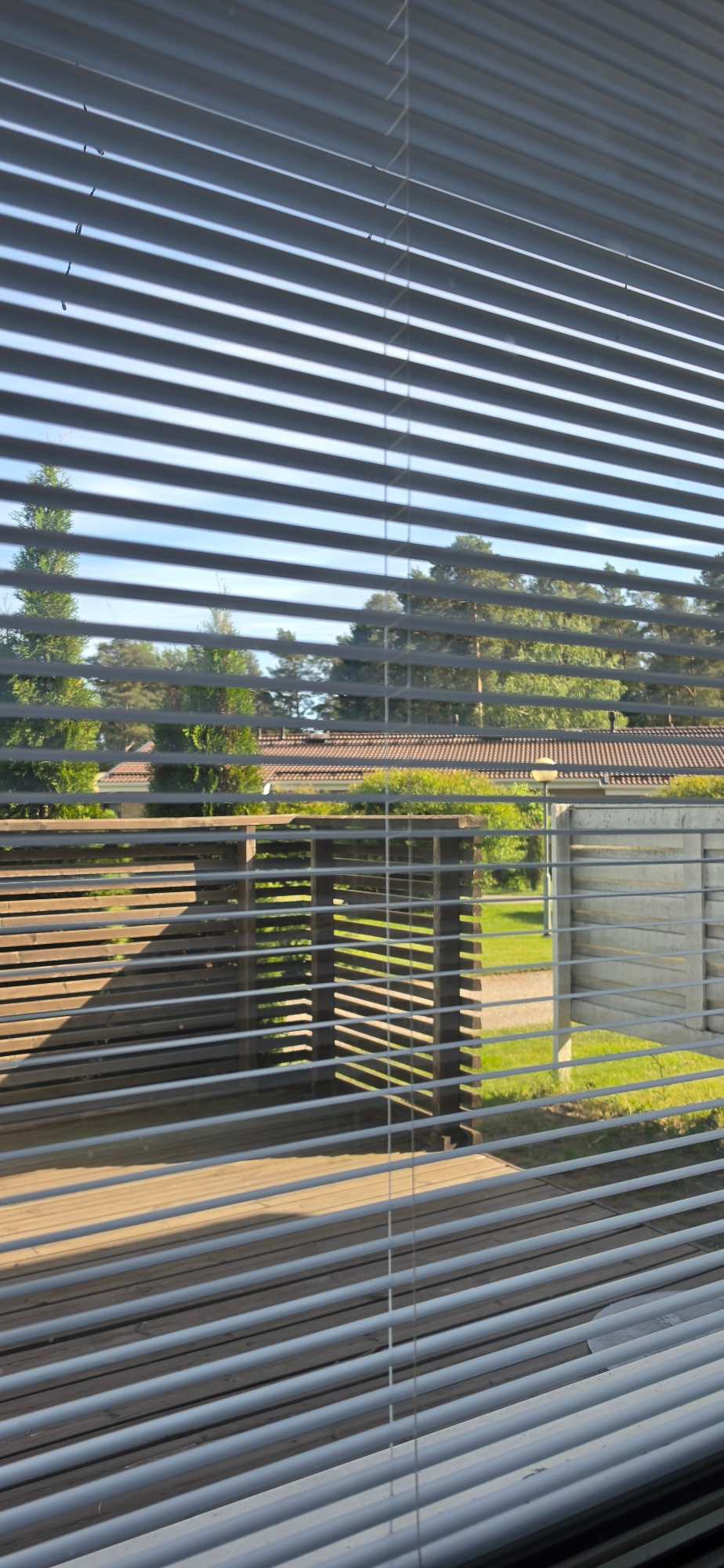 The image depicts a view through horizontal blinds onto a backyard scene in Uusikaupunki, Finland. The blinds are silver and cast striped shadows on a wooden deck in the foreground. Beyond the deck, there is a wooden structure with slatted sides, a paved pathway, and a patch of green grass. In the middle ground, a light-colored concrete fence with horizontal slats is visible, and behind that, a building with a red-tiled roof and some trees. The background features a clear blue sky and more trees, consistent with a residential area in Finland. There are no people visible in the image. The overall impression is a sunny day with the blinds partially obscuring the view.