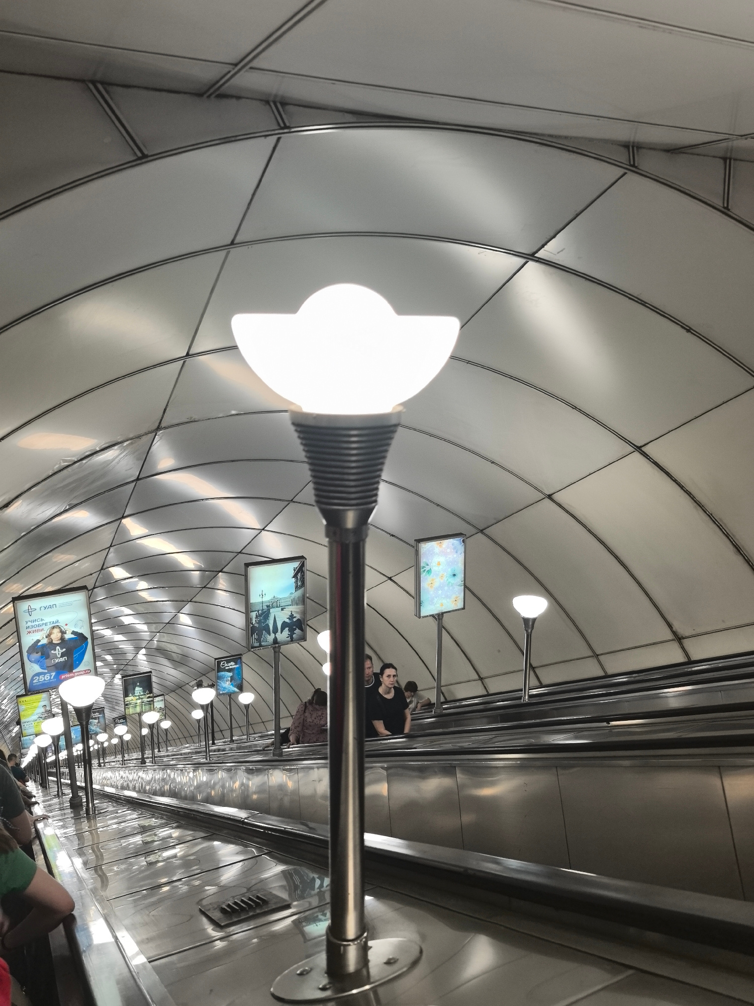 In Saint Petersburg, Russia, this image captures an upward-facing escalator in a metro station. The ceiling is a striking feature, composed of curved, metallic panels arranged in a repeating pattern, with visible structural beams. Along the escalator's sides, illuminated advertising displays are mounted on poles, some featuring text in Cyrillic script. Several people are present, some on the escalator and others standing near the top. The escalator itself is made of polished metal, reflecting the ambient light. A prominent light fixture, with a distinctive white, petal-shaped shade, stands in the foreground, its metallic pole extending upwards. The overall environment is modern and functional, characteristic of a well-lit urban transportation hub.