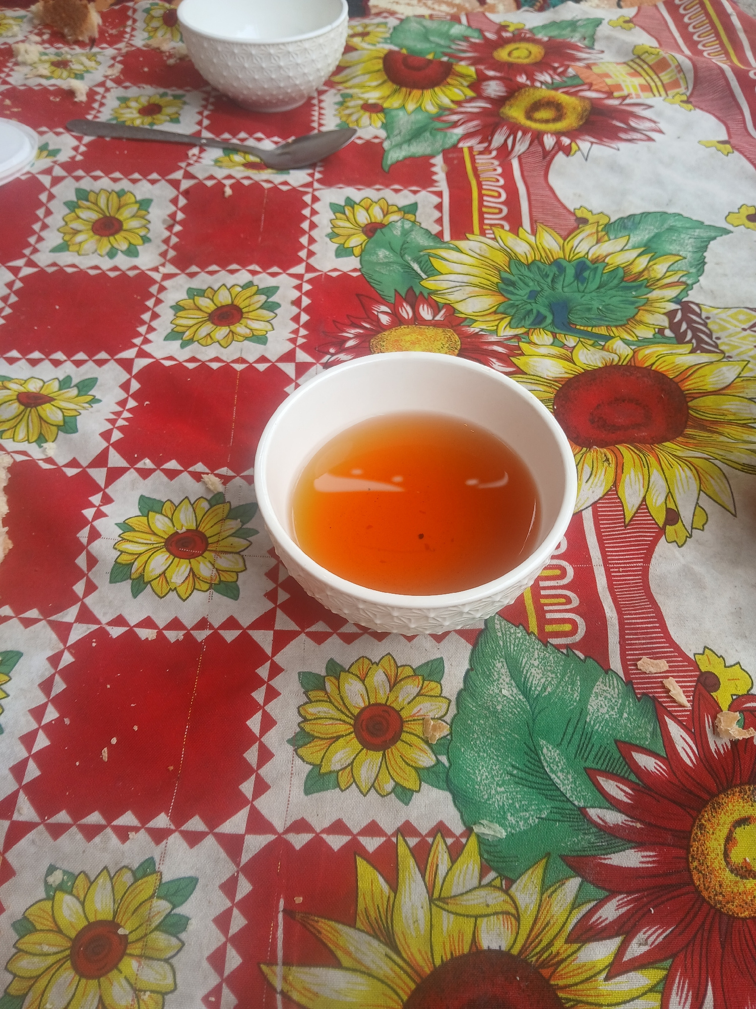 The image depicts a close-up view of a table covered with a tablecloth featuring a pattern of sunflowers and a red and white checkered border. A white bowl filled with amber-colored liquid, likely tea, is placed in the center of the frame. To the left of the bowl, a metal spoon rests on the tablecloth. Another white bowl, empty, is visible in the upper left corner. The scene appears to be indoors, possibly a dining area, and there are small crumbs scattered across the tablecloth. The image does not provide information about the city of Kotormo, Kyrgyzstan, or any specific location within it.