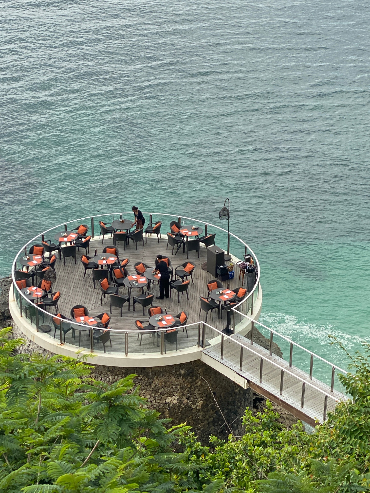 The image shows an outdoor restaurant with a circular dining deck overlooking the ocean. The deck is made of wood and features numerous small tables with orange-cushioned chairs. Two staff members, likely waiters, are visible on the deck, one attending to a table and the other in motion. A ramp with metal railings connects the deck to a pathway, suggesting it's part of a resort or cliffside establishment. The ocean water is a vibrant turquoise, with gentle waves breaking against the rocky shore below. Lush green foliage frames the bottom of the image, indicating a tropical setting. The restaurant's location is likely in Kuta Selatan, Indonesia, known for its scenic coastal views and upscale dining establishments.