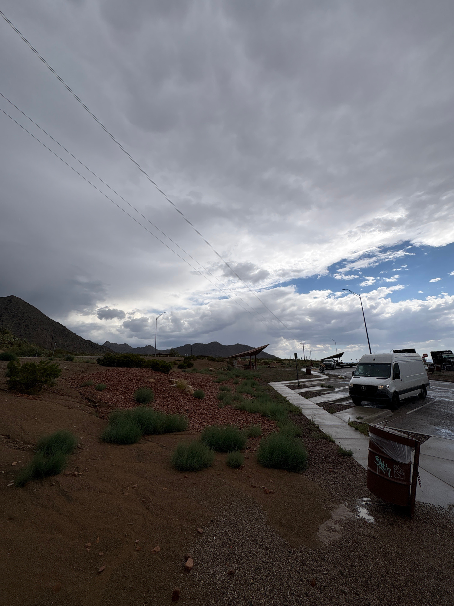 The image captures a roadside scene with a dark, overcast sky and scattered clouds, suggesting recent or impending rain. In the foreground, a dirt and gravel embankment is dotted with low-lying green vegetation and reddish-brown rocks. A paved pathway runs along the side of the road, which appears wet, indicating rain.
Further into the scene, a white cargo van is parked on the right side of the wet pavement. A brown trash receptacle with visible graffiti and a plastic liner is positioned near the edge of the pavement. Across the road, several vehicles, including what appears to be a larger truck, are visible. In the background, there are mountains and a covered picnic area with a distinctive triangular roof structure. Overhead, power lines are strung across the frame. The general location appears to be a rural or semi-rural area. There are no people visible in the image.