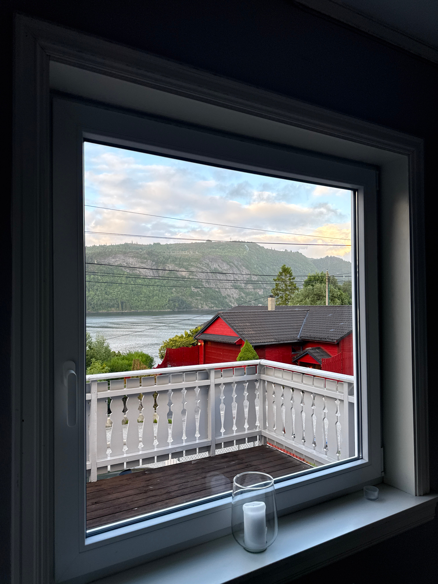 A view from inside a building, through a white-framed window, looking out onto a balcony and a natural landscape. The balcony is constructed with wooden decking and a white railing featuring an ornate, decorative design with alternating upright posts and open spaces. A glass candle holder with a white pillar candle sits on the white windowsill.
Beyond the balcony, a red building with a dark roof is partially visible. The landscape consists of a body of water, likely a fjord, with a densely forested mountainside rising behind it. Power lines crisscross the scene, strung across the mountains. The sky above is partially cloudy, with soft, diffused light suggesting either early morning or late evening. The scene is tranquil and appears to be a residential area, possibly in or near Bergen, Norway, given the characteristic fjord and mountainous terrain. There are no people visible in the image.