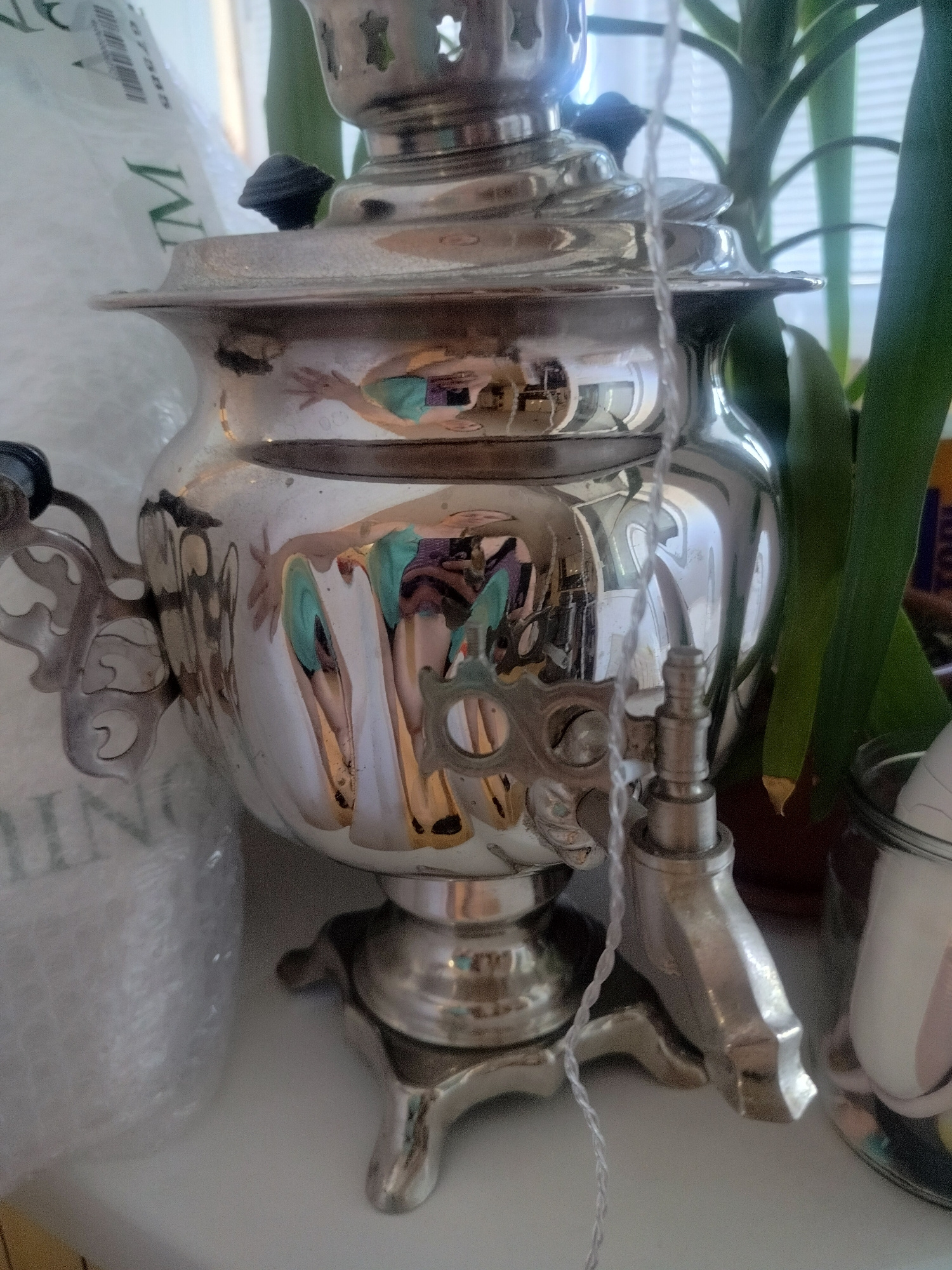 A close-up, medium shot captures a highly polished, silver-colored samovar positioned on a white surface. The samovar exhibits ornate detailing and has a spigot on its right side. A thin, white string is draped across the front of the samovar. The background is a shallow depth of field, revealing a portion of a large green plant with broad leaves to the right, and a white, translucent, crinkled material on the left, possibly bubble wrap. The surface the samovar rests on appears to be a windowsill, with a window and blinds visible in the upper background. Reflections are prominent on the polished surface of the samovar, showing distorted images of what appear to be people wearing colorful clothing. The image lacks any specific indicators of its location in Novosibirsk, Russia.