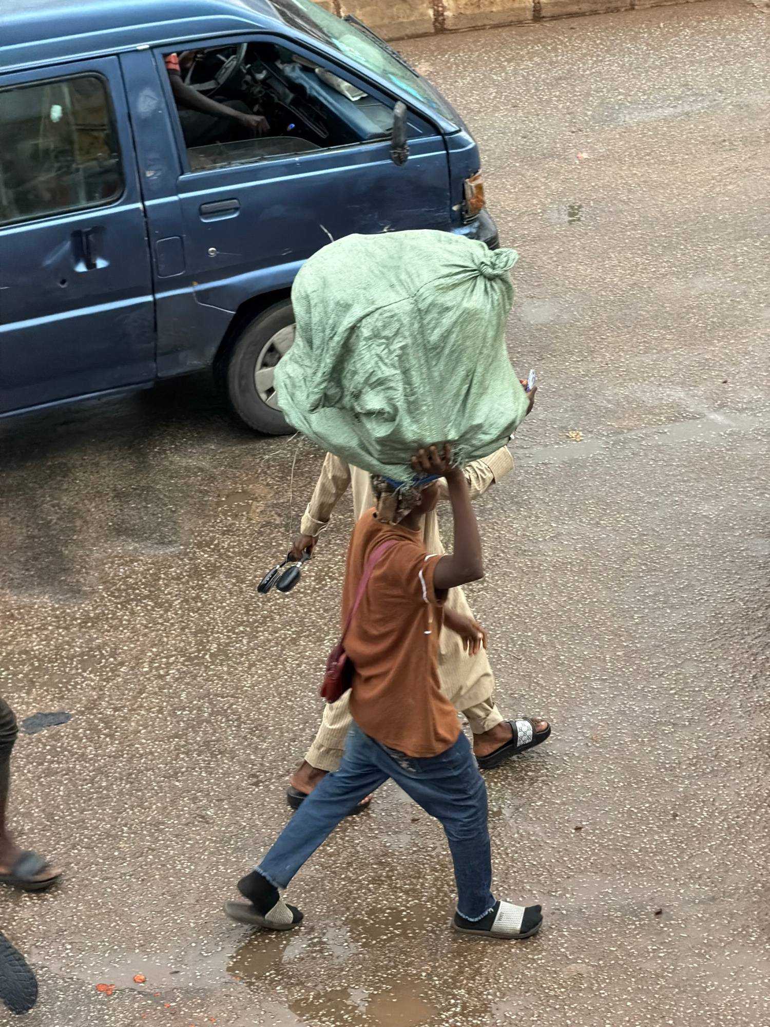 In Bukuru, Nigeria, a man in a brown shirt and blue jeans walks across a wet, speckled asphalt surface, carrying a large, light green, knotted bag over his head. He wears black socks and patterned slide sandals. To his right, another person in a tan robe walks in the same direction, holding a pair of black objects that appear to be folded scissors or similar tools. A blue minivan is parked to the left of the frame, its driver's arm visible inside. The ground shows reflections from the wetness.