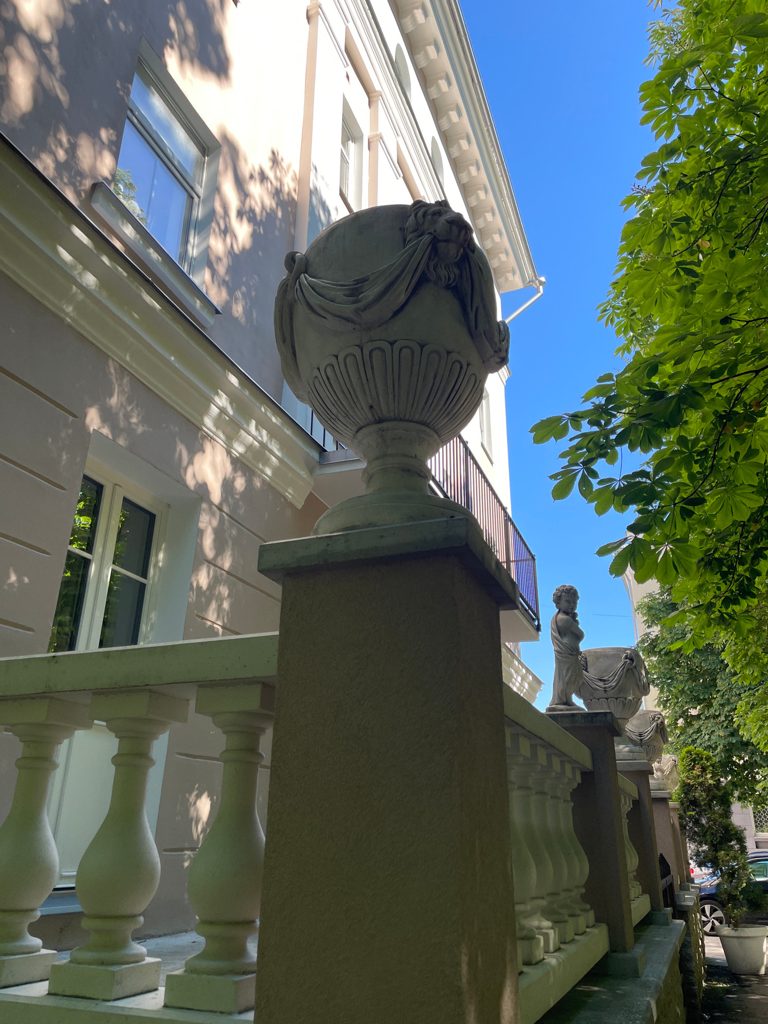 The image depicts a low-angle view looking upwards towards a building with classical architectural elements, under a clear blue sky and dappled sunlight filtering through green foliage from overhead trees.
The primary subjects are architectural features: a large, ornate stone urn or finial prominently positioned in the foreground, adorned with intricate carvings. Behind it, a balustrade with classical spindles runs along a staircase or balcony. The building itself appears to be a light-colored structure with visible windows. Further in the background, a statue of a human figure can be seen, also in a classical style, standing on a higher level. The overall aesthetic suggests a historical or traditionally designed building.
The scene appears to be set outdoors during the daytime, given the bright sunlight and blue sky. The presence of leaves indicates it is likely spring or summer. The location context provided places this in Tallinn, Estonia, which might imply a European city setting with such architectural styles. There is no discernible activity or event taking place; the image is a still, architectural study. Notable details include the strong interplay of light and shadow on the building's facade, cast by the overhead trees, and the clear, unblemished blue sky. The visual composition emphasizes the grandeur and decorative nature of the architecture. No text is visible in the image.