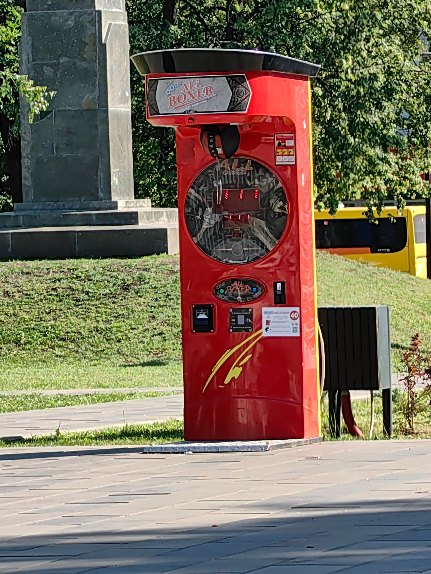 A red, vertically oriented boxing arcade game, branded "ALI BOXER PRODUCTION," stands outdoors. The game features a large circular screen showing the interior mechanism and lights. Above the screen, a graphic with the "ALI BOXER" logo is visible. Below the screen, there are various control panels and displays, including a coin slot and a digital readout showing "40". A yellow and black object, possibly a bus or public transport, is visible in the background, partially obscured by lush green trees and a grassy slope. To the left of the boxing machine, a large, gray, stone monument with steps is situated. The scene appears to be in a park or public square, suggested by the paved walkway in the foreground and the manicured grass. The time of day is likely daytime, indicated by the bright sunlight casting shadows. The weather appears to be clear. There is no indication of people or any specific activity taking place, beyond the potential for use of the boxing game.