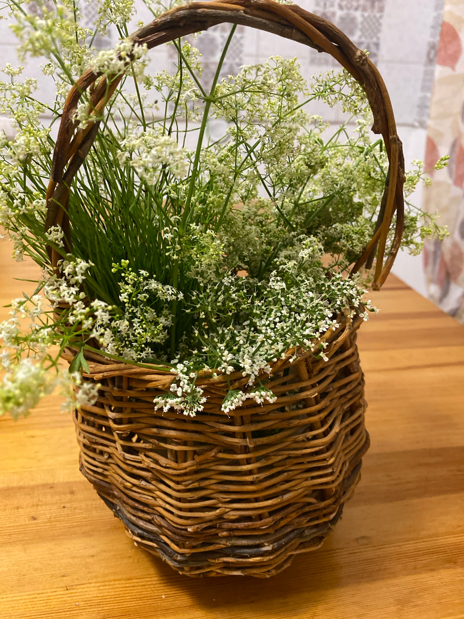 A rustic, woven wicker basket overflows with delicate white flowers and greenery, placed on a polished wooden table. The basket has a curved handle made of twisted branches. The background features a blurred pattern of tiles and curtains, suggesting an indoor setting. The image is a close-up, with the basket filling most of the frame. The scene is static and appears to be a decorative arrangement. The location is Zelenodol'sk, Russia.
