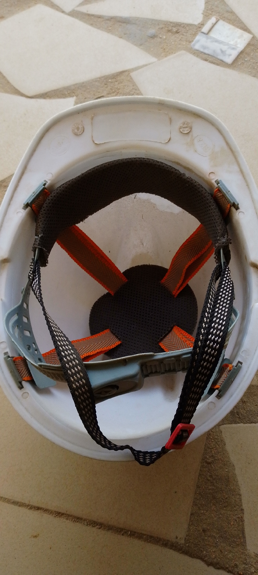 A white construction hard hat is shown from an overhead perspective. The interior of the hard hat is visible, revealing a suspension system with straps and padding. The hard hat rests on a textured surface that appears to be made of large, irregularly shaped tiles or stones in various shades of tan and beige. Some of these tiles are broken or chipped. The lighting suggests an outdoor environment, possibly during daylight hours, with the sun casting subtle shadows. There are no people or animals present in the image, and the scene appears to be static and undisturbed. The hard hat is the central focus of the image, with its internal components clearly discernible. The overall impression is one of an object left on a ground surface.