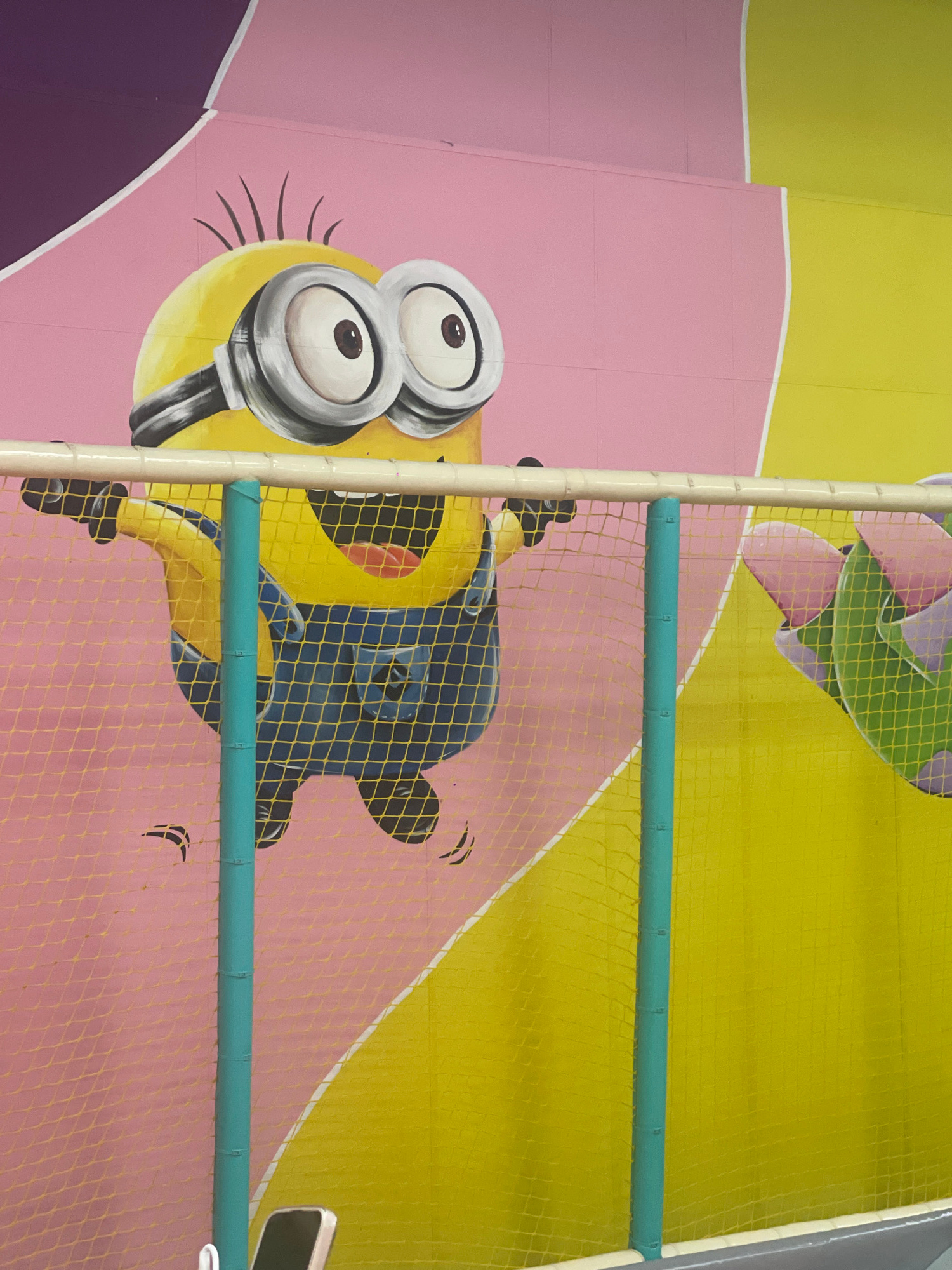 The image is a photograph of a colorful mural featuring a Minion character painted on a wall. The Minion is depicted in its typical yellow form, wearing blue overalls and goggles, with a wide smile and its arms outstretched. The wall behind the Minion is painted with large blocks of pink, yellow, and purple, with some white lines creating a geometric pattern. A yellow net barrier with turquoise posts is in the foreground, partially obscuring the lower portion of the Minion and the wall. The environment appears to be an indoor play area, possibly a playground or a children's entertainment center. The image does not provide specific information about the city of Pavlodar, Kazakhstan.