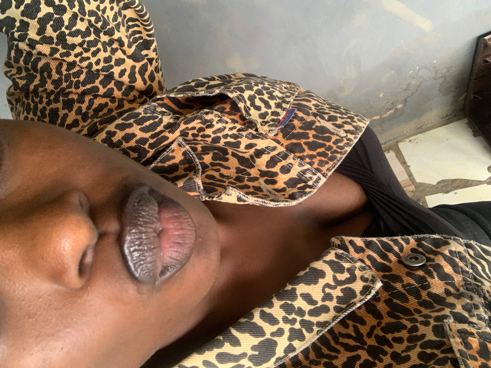 A close-up, high-angle shot captures a person's face and upper body, with a focus on their lips and nose. The individual is wearing a leopard-print jacket over a black top. The jacket features visible stitching and a button closure. The background is a neutral, grey wall with some discoloration and a tiled floor. The scene appears to be indoors, possibly in a residential or public space within New Stadium, Nigeria.
