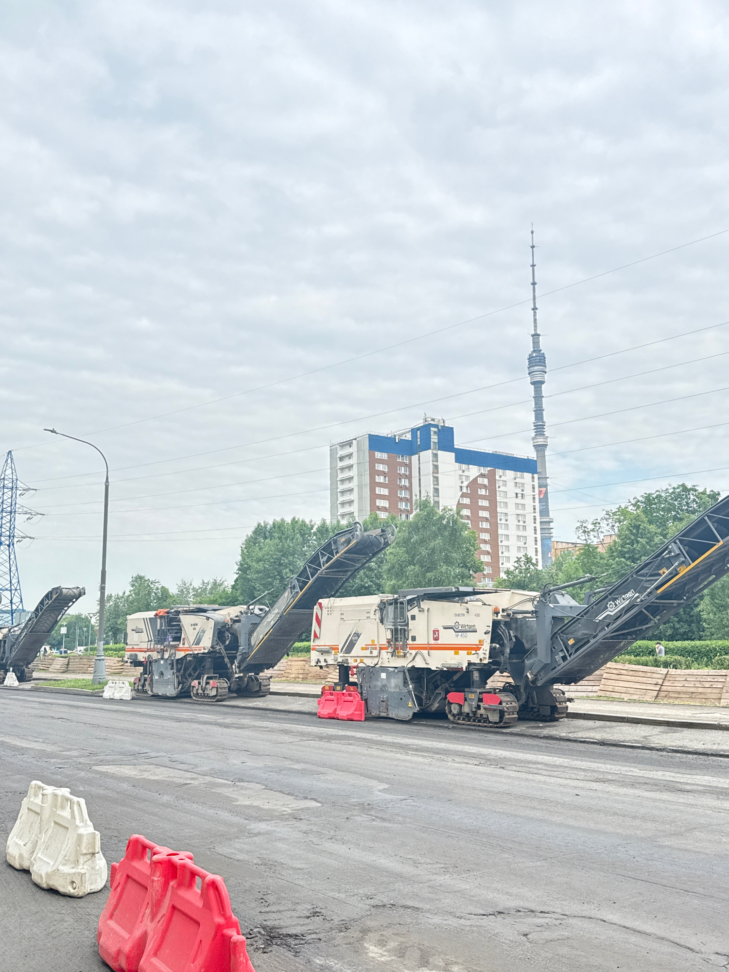 The image depicts a construction site on a cloudy day in Moscow, Russia, likely on an overcast summer day, with a view of the Ostankino Tower in the background. In the foreground, a partially paved road shows signs of recent work, with visible patches of asphalt and some minor damage. Several pieces of heavy construction equipment, specifically milling machines, are positioned on the road, indicating road resurfacing or repair.
Two white and red plastic barriers are placed on the road in the foreground, serving as temporary traffic control. Further down the road, two milling machines are visible. The milling machines are large, industrial vehicles with conveyor belts and rotating drums designed to remove existing pavement. The one in the foreground has "WIRTGEN" branding. Red plastic barriers are also placed near the milling machines.
In the middle ground, behind the construction equipment, there is a row of lush green trees. Beyond the trees, a modern apartment building with white and blue facades stands tall. A tall, slender lattice tower, the Ostankino Tower, is visible in the distance, piercing the overcast sky. A power transmission tower is also present on the left side of the image, adding to the urban industrial landscape. The overall scene suggests ongoing road construction and infrastructure maintenance within a populated urban area of Moscow.