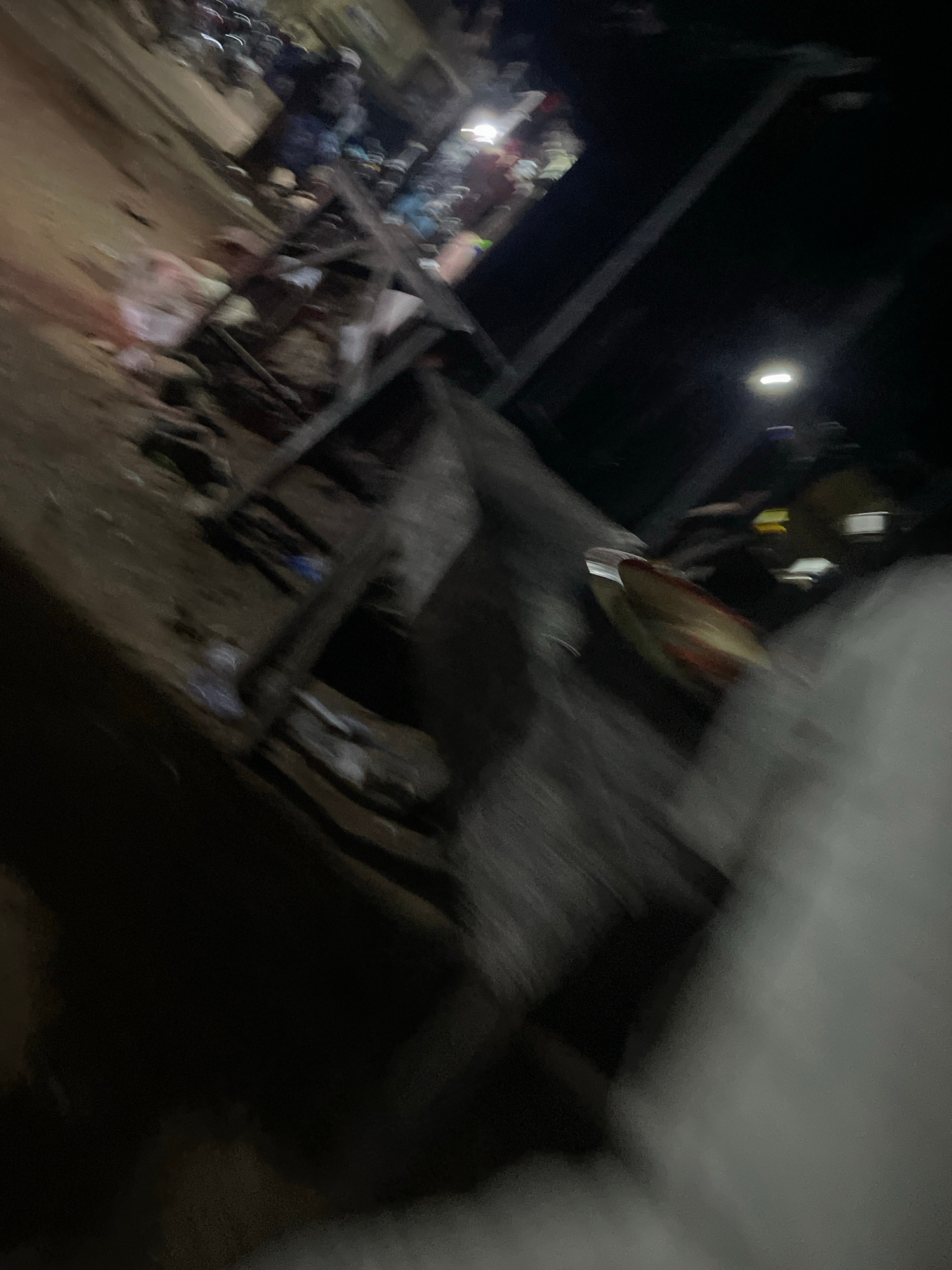 The image depicts a chaotic scene, likely in a market or a busy public area in Lokoja, Nigeria. The foreground is dominated by blurred figures and debris, suggesting a dynamic environment with movement. In the midground, there are structures that appear to be stalls or temporary shelters, with various objects strewn around. A closer look reveals what might be people gathered, though their features are indistinct due to motion blur and low lighting. Some appear to be wearing dark clothing, while others have lighter colored attire. There are also hints of what could be food items or merchandise on display or in containers. The background is largely dark, but there are indications of streetlights or artificial lighting, casting a glow on some areas and adding to the overall sense of activity. The overall impression is one of a bustling, possibly crowded, evening scene in an urban setting.