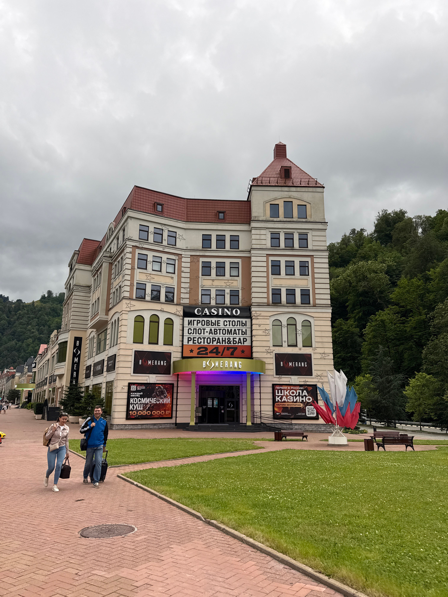 The image shows a multi-story building in the city of Estosadok, Russia, with a red tiled roof and beige facade. The building appears to be a casino and entertainment complex, indicated by prominent signage including "CASINO," "IGROVYE STOLY" (game tables), "SLOT-AVTOMATY" (slot machines), "RESTORAN & BAR," and "SHKOLA KAZINO" (Casino School). There are also advertisements for "KOSMICHESKIY KUSH" (Cosmic Prize) with a monetary amount.
In the foreground, a paved brick walkway leads towards the building. Two people are walking on the walkway away from the camera. A woman on the left is wearing a light pink jacket and blue jeans, carrying a brown bag over her shoulder and a larger brown tote bag in her hand. A man on the right is wearing a blue jacket and blue jeans, pulling a black rolling suitcase. They are walking side-by-side, appearing to interact casually.
To the right of the walkway is a well-maintained green lawn with some scattered white flowers. A pathway of grass separates the lawn from the walkway. Further back, near the entrance to the casino, are benches and a trash receptacle. In front of the building, a cluster of red, white, and blue flags on a stand are visible.
Behind the building, a dense forest of green trees covers a hillside. The sky is overcast with gray clouds. The overall atmosphere suggests an urban or resort setting on a cloudy day.