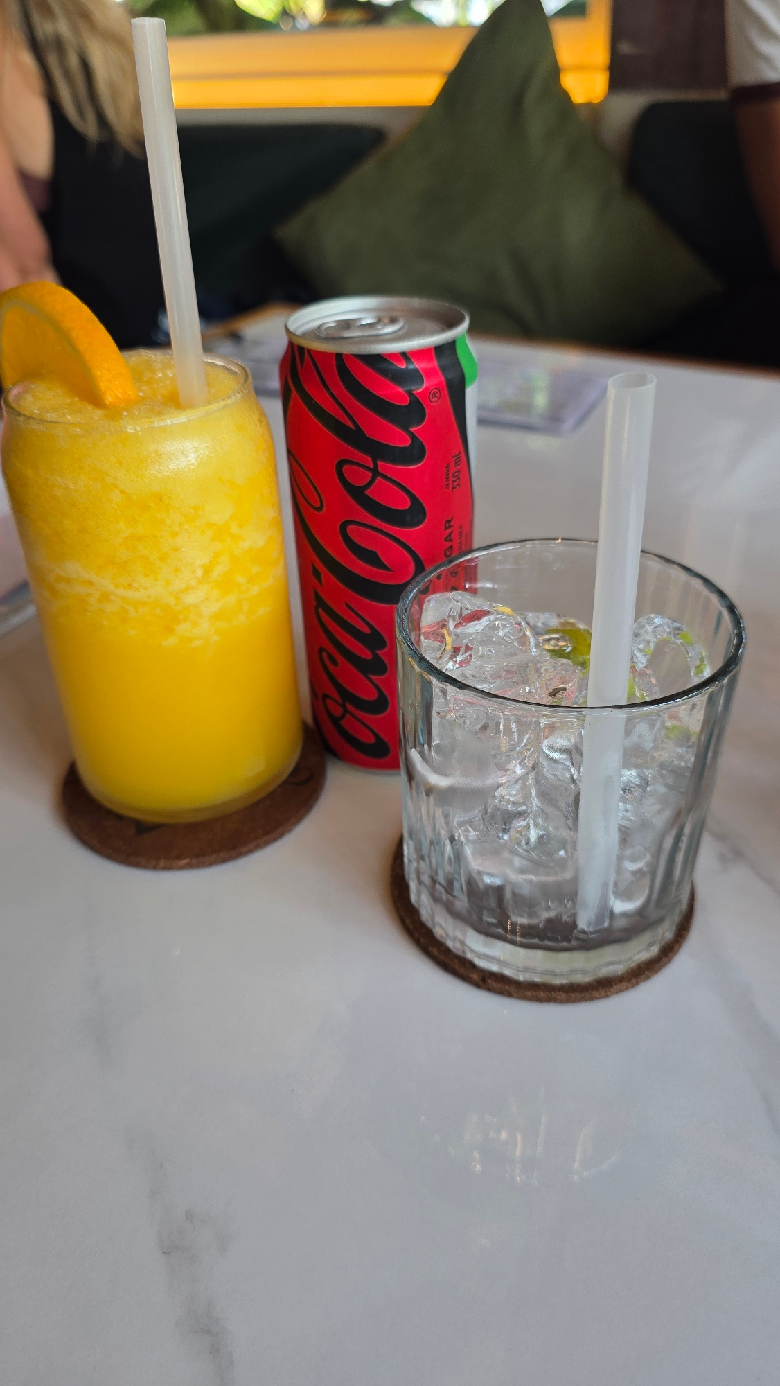 A close-up, eye-level shot captures a table setting with three drinks in a restaurant. On the left, a tall glass contains a bright orange, frothy beverage, likely a smoothie or juice, garnished with an orange slice and a white straw. To its right, a red and black can of Coca-Cola Zero Sugar stands prominently. Next to the can, a clear glass is filled with ice and a clear liquid, possibly water or a cocktail, with a white straw inserted. The drinks are placed on a light-colored, marble-patterned table with round, brown coasters underneath the glass and can. In the blurred background, a person with blonde hair is visible wearing a dark top, and a green cushion is on a sofa. The overall lighting suggests an indoor setting. The image does not provide specific information about the city of Mengwi, Indonesia.