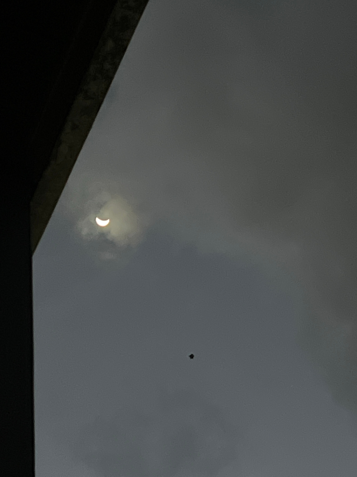 An upward-facing, medium-angle shot captures a cloudy sky from a dark, overhanging structure. A crescent-shaped sun, partially obscured by a cloud, is visible in the upper left quadrant of the frame. Below and to the right of the sun, a small, dark, kite-like object floats in the sky. The sky is overcast with varying shades of gray clouds.